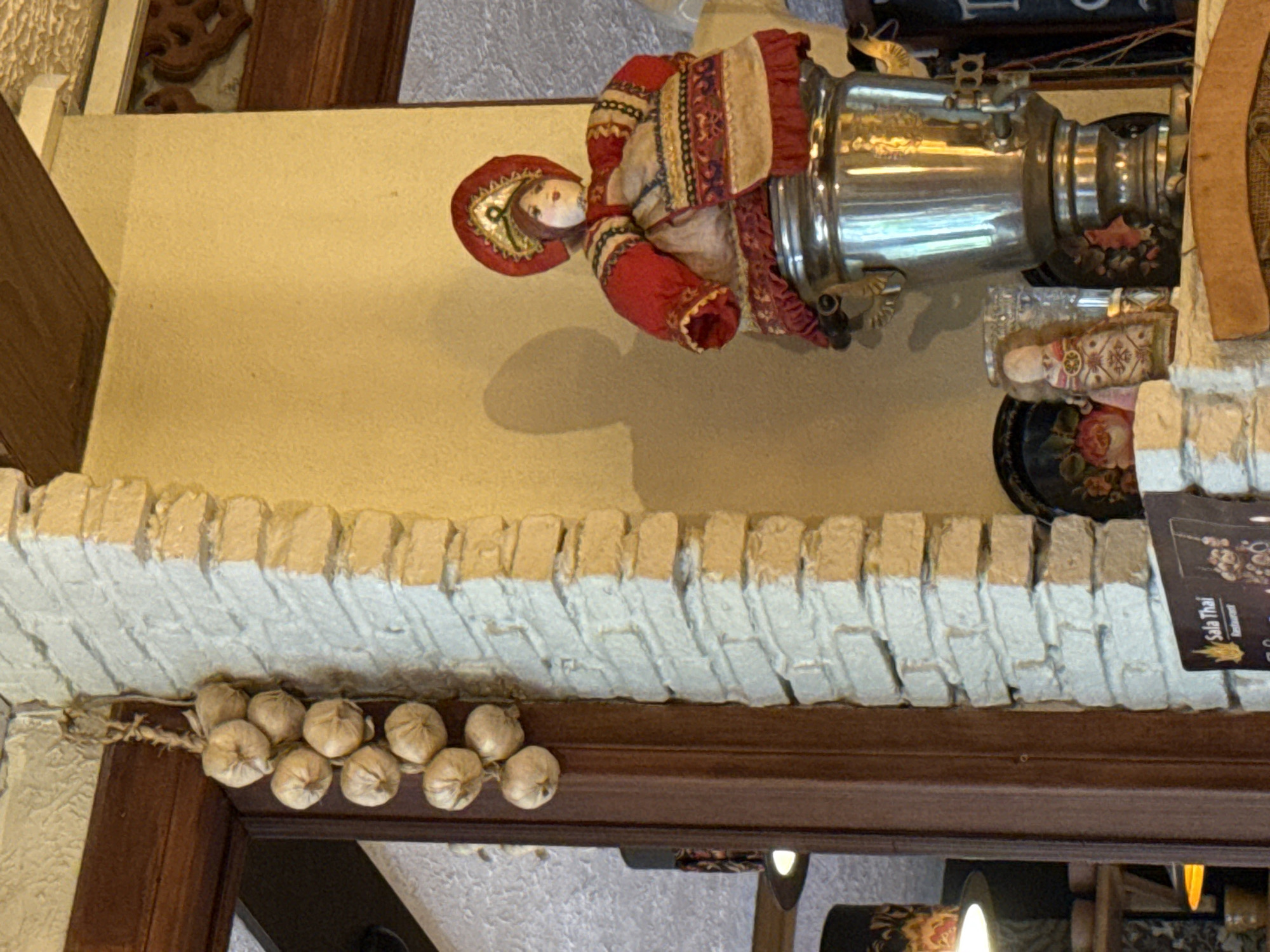 Indoor scene in Baan Chaweng Noi, Thailand, featuring a decorative Russian doll with a samovar, garlic string hanging from a brick ledge with a sign reading "Sala Thai Restaurant" and a partially visible wooden decorative panel.