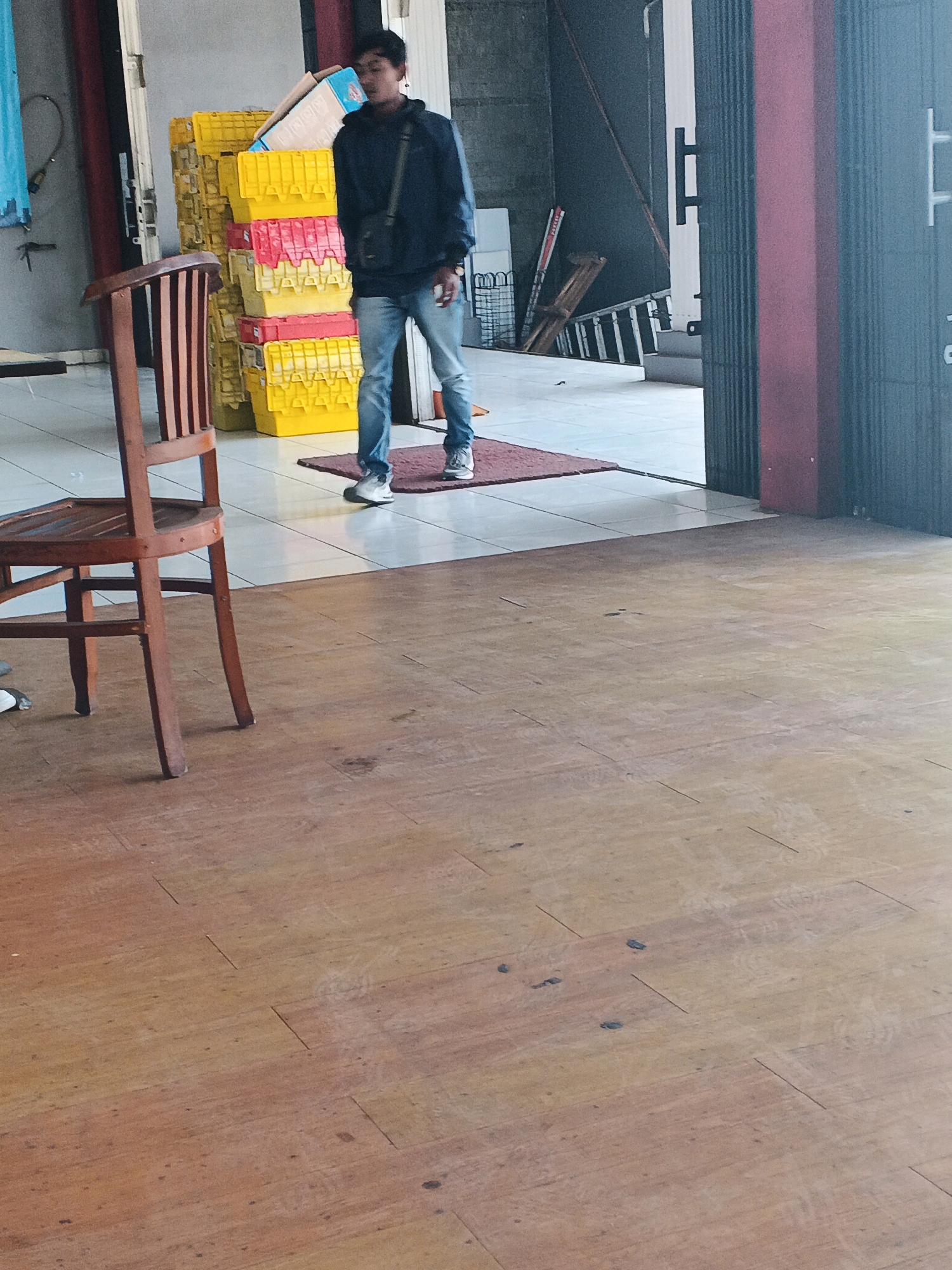 A young man stands in front of a stack of yellow and red crates. He is wearing a dark blue jacket and jeans. He is looking down and to his left. The man is standing on a red rug in front of the crates. A wooden chair is in the foreground to the left of the man. The floor is made of wood planks. The background shows a building with a dark gray wall and a red pillar. There are also some ladders and other objects in the background. The location is Genuk Barat, Indonesia.