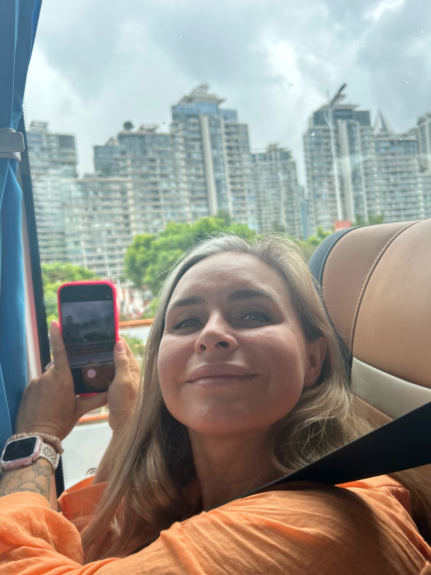 A woman with blonde hair and blue eyes is taking a selfie inside a vehicle, possibly a bus or car, with her phone. She is wearing an orange top and a watch on her left wrist. The background shows a cityscape with tall buildings and trees, indicative of a metropolitan area like Jing'an District, China. The sky is overcast. A seatbelt is visible across her shoulder.