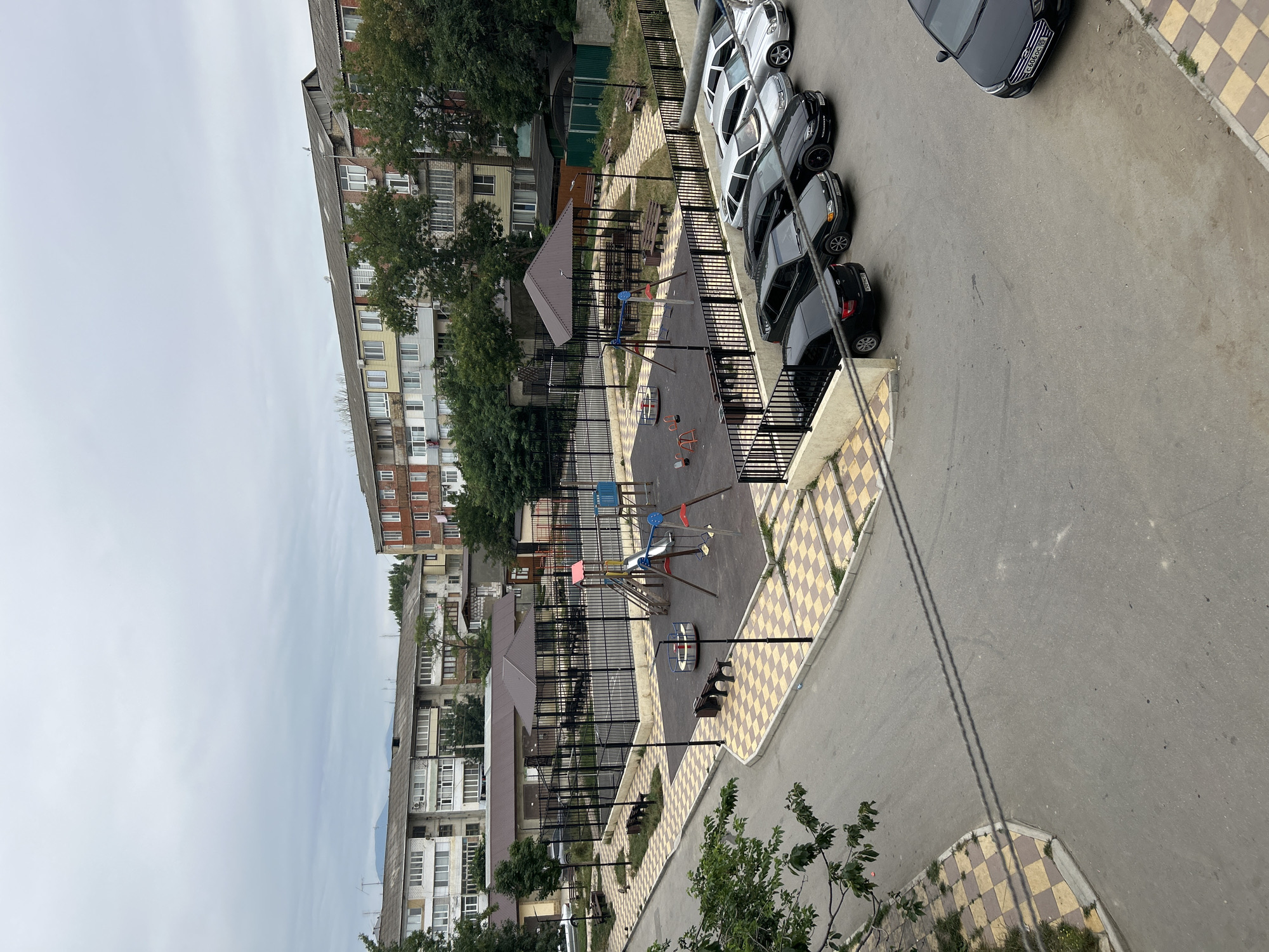 This is an aerial, eye-level, medium shot capturing a residential courtyard in the city of Buynaksk, Russia, on an overcast day. The scene is dominated by apartment buildings, a playground, and a parking lot filled with cars.
The apartment buildings are typical of Soviet-era architecture, with multiple stories, uniform windows, and balconies. They appear to be in varying states of repair, with some showing signs of age and others appearing more recently renovated with new siding or paint. The buildings are arranged in a U-shape around the courtyard, creating a sense of enclosure.
In the center of the courtyard is a fenced-in playground with swings, a slide, and a merry-go-round. The playground is surrounded by a rubberized safety surface. Adjacent to the playground is a covered seating area with benches.
A paved road curves through the courtyard, with cars parked along its edges. The cars are a mix of makes and models, and most appear to be sedans. The road is lined with sidewalks and a checkerboard pattern of yellow and beige tiles.
The environment is somewhat urban and densely populated, as evidenced by the number of apartment buildings and cars. The overcast sky suggests a cooler climate, and the presence of trees indicates a temperate region. There are no people visible in the image.
