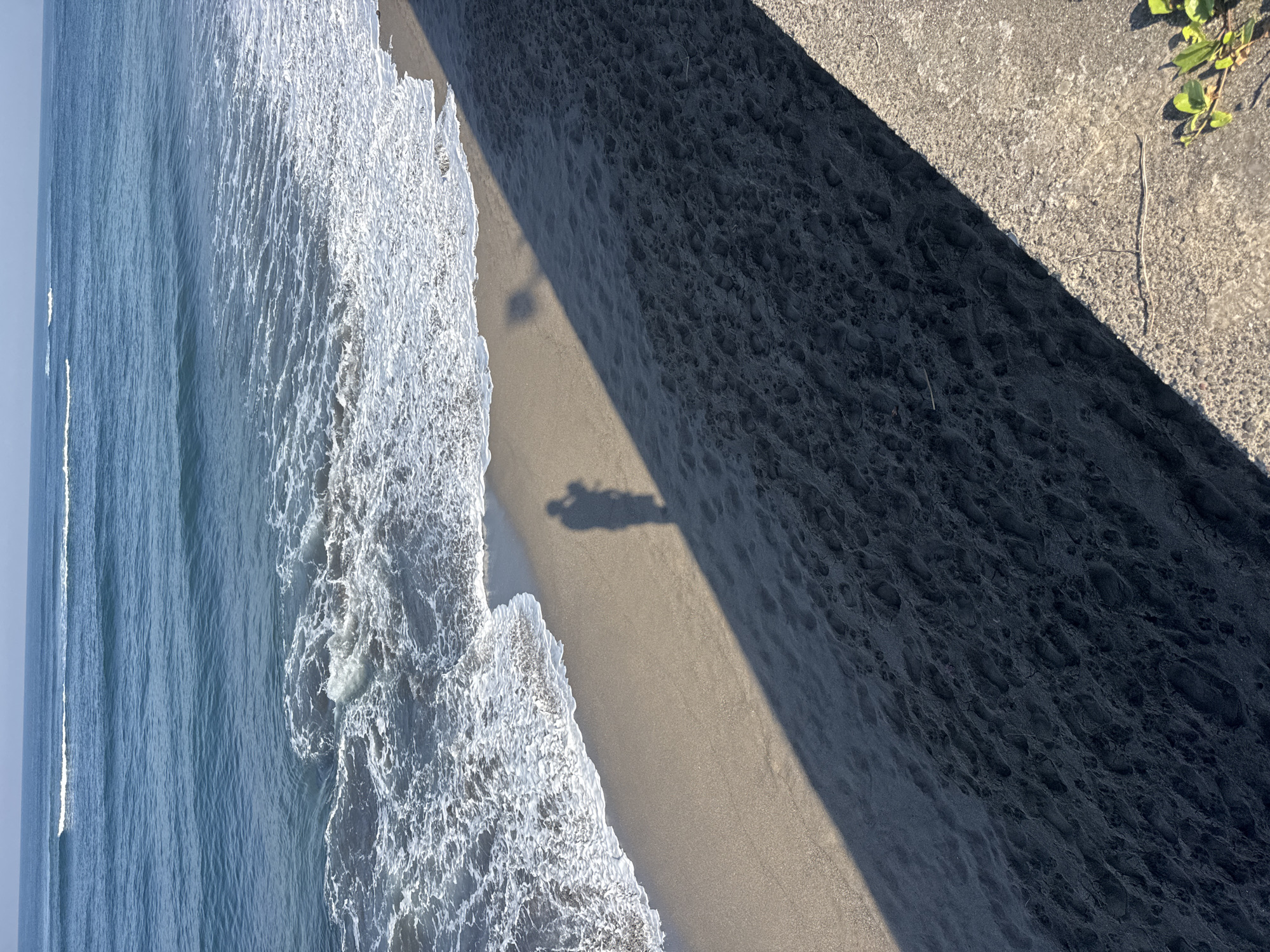The image displays an elevated, downward-looking view of a sunlit coastline, likely in Mengwi, Indonesia. The left side is dominated by the ocean, with blue-green water extending towards the horizon under a bright sky. White, foamy waves are seen breaking and receding along the shore, creating a dynamic texture where they meet the sand.
To the right, a sandy beach curves along the water's edge. A sharp, diagonal shadow, cast by an unseen elevated structure or cliff, stretches across a significant portion of the sand. The section of the beach in direct sunlight is a light, sandy brown. On this brightly lit sand, near the breaking waves, a distinct, elongated shadow of a person, possibly holding a device like a camera or phone, is visible. This indicates the photographer is standing on the elevated platform.
The part of the beach enveloped by the large shadow appears darker, showing subtle textures and disturbances, possibly footprints. In the upper right corner, the edge of a light-colored concrete or stone surface is visible, brightly illuminated by the sun, from which the perspective originates. Small green foliage can be seen growing near this edge.
The long shadows suggest the time of day is either early morning or late afternoon. The scene conveys a bright, clear day by the coast. No visible text is present in the image.