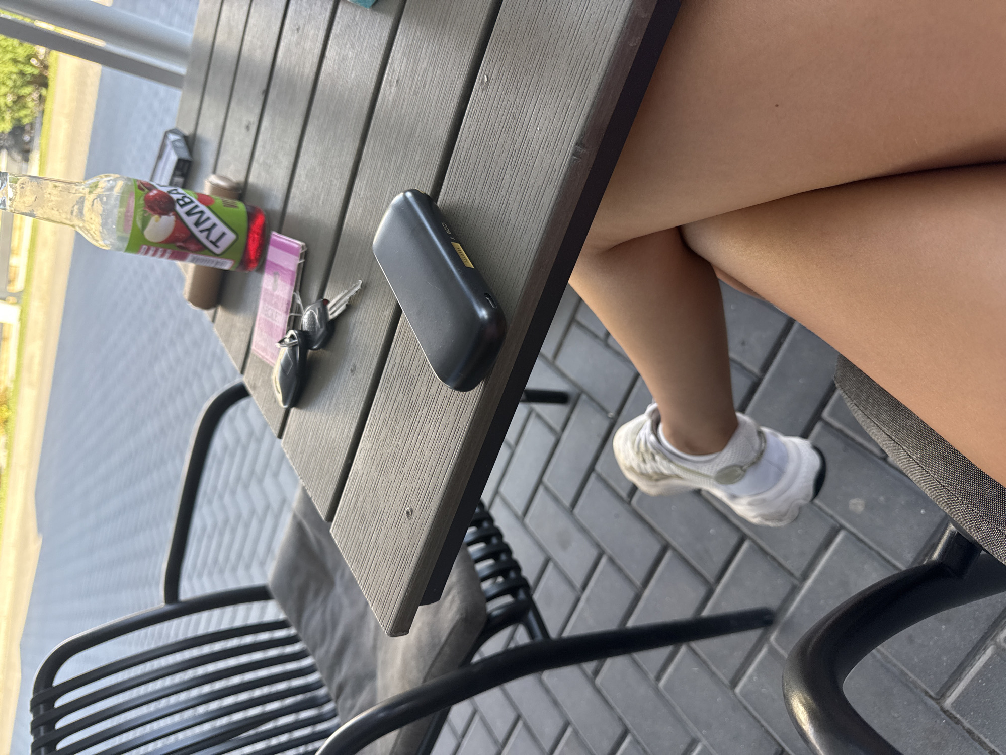 The image depicts a close-up of a person sitting at an outdoor table. The scene appears to be an outdoor cafe or seating area, as indicated by the chairs, table, and paving stones. On the table, there is a glass bottle with a label that reads "TYMBA" and appears to contain a red liquid, possibly a beverage. Also visible on the table are a pack of cigarettes, a pair of car keys, and a black electronic device. The person's legs and feet are visible in the foreground, with one foot wearing a white sneaker. The overall setting is outdoors, with a paved ground surface. The location is specified as the city of Comrat, Moldova, implying this is a scene within that city.