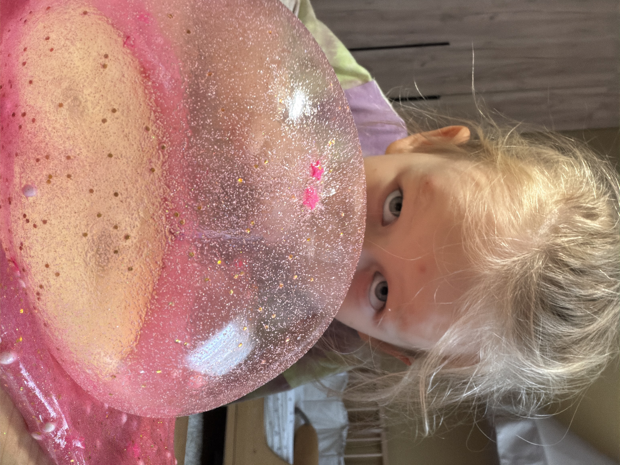 A young Caucasian girl with blonde hair is looking over a large pink, glitter-filled bubble. The bubble is translucent and appears to be made of slime or a similar substance. The girl's face is partially obscured by the bubble, but her eyes are visible, looking directly at the camera. She is wearing a tie-dye shirt with purple and green colors. The background is slightly blurred, showing a wooden cabinet on the left and what appears to be a crib or bed with a railing on the right. The overall lighting suggests an indoor setting. No specific location in Khimki, Russia is identifiable from the image.