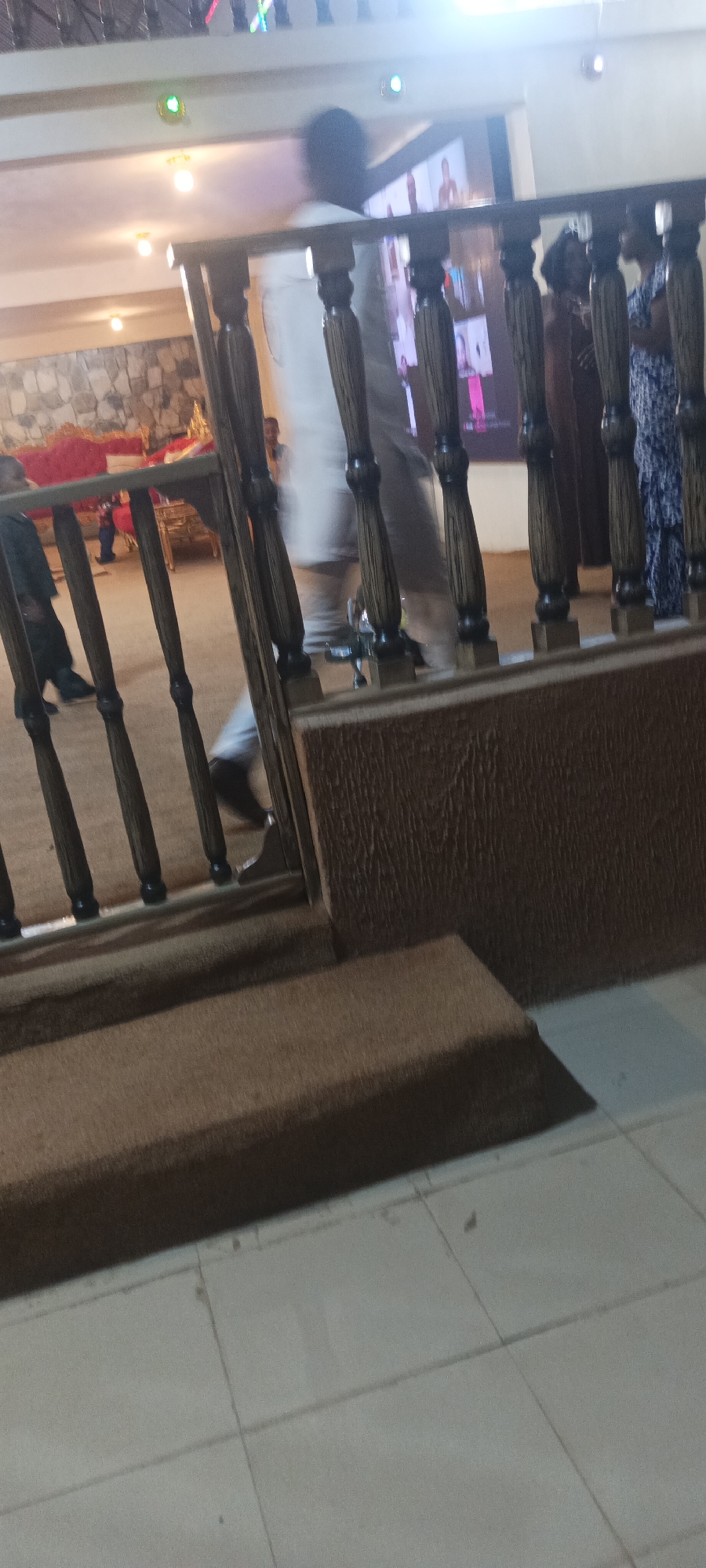 The image shows a blurred interior scene, likely a venue or event space. A wooden railing with turned balusters is prominent in the foreground, obscuring much of the background. Beyond the railing, several people are visible, but their features are indistinct due to motion blur or low resolution. One person in a white garment is walking, and others are standing or interacting. A large screen displaying images of people is visible in the upper background. A stone wall and a red upholstered bench are also partially visible. The floor appears to be tiled. The scene is lit with overhead lighting. This image does not provide any specific information about the location being on Rukuba Road, Nigeria.