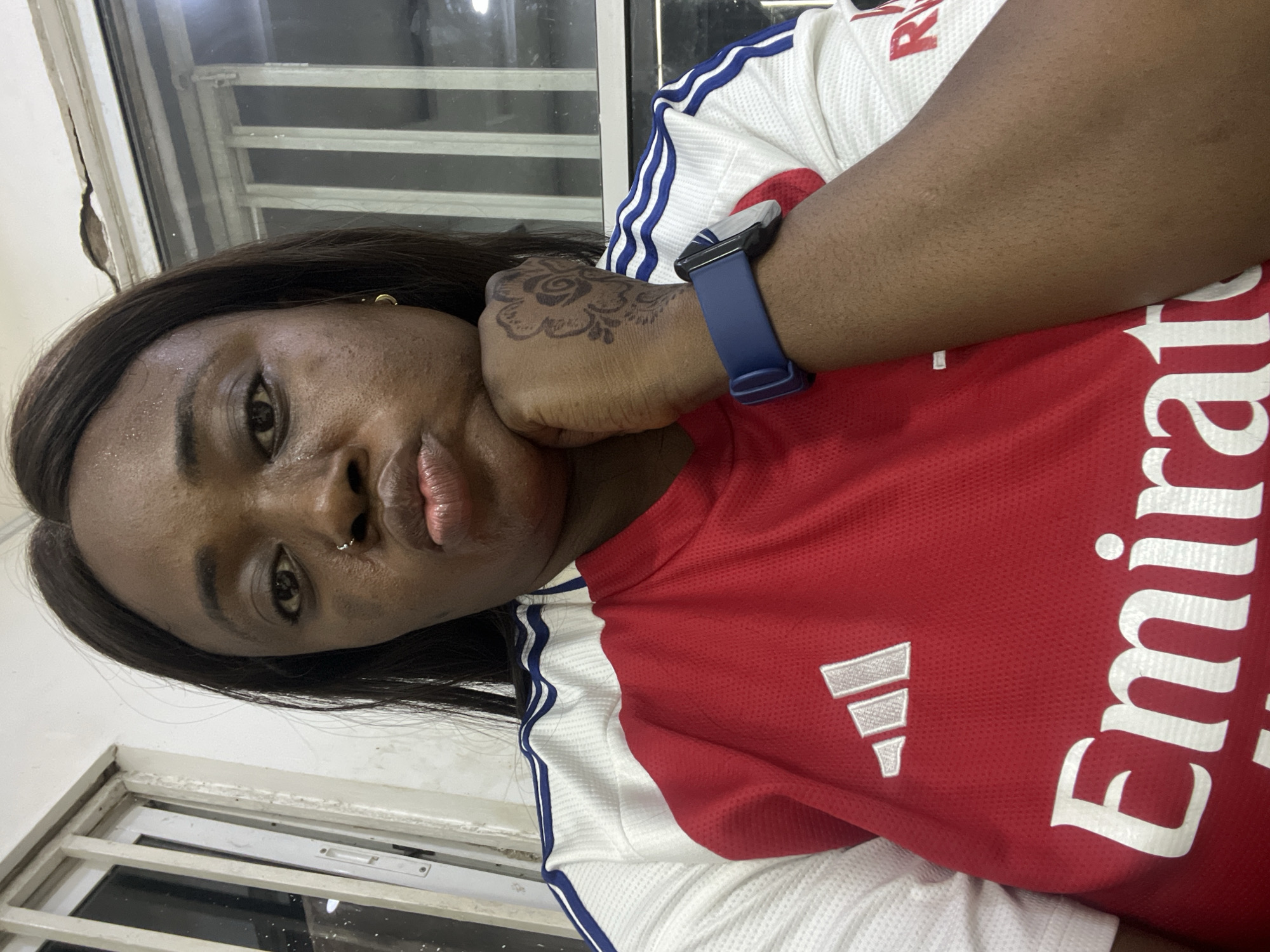 A woman is pictured indoors, her face angled towards the camera. She has dark hair, dark eyes, and a nose piercing. She rests her head on her hand, which is adorned with a tattoo. She wears a red and white soccer jersey with "Emirates" printed on it. A blue smartwatch is on her wrist. A window frame is visible in the background. The location in Kubwa, Nigeria cannot be determined from the image.