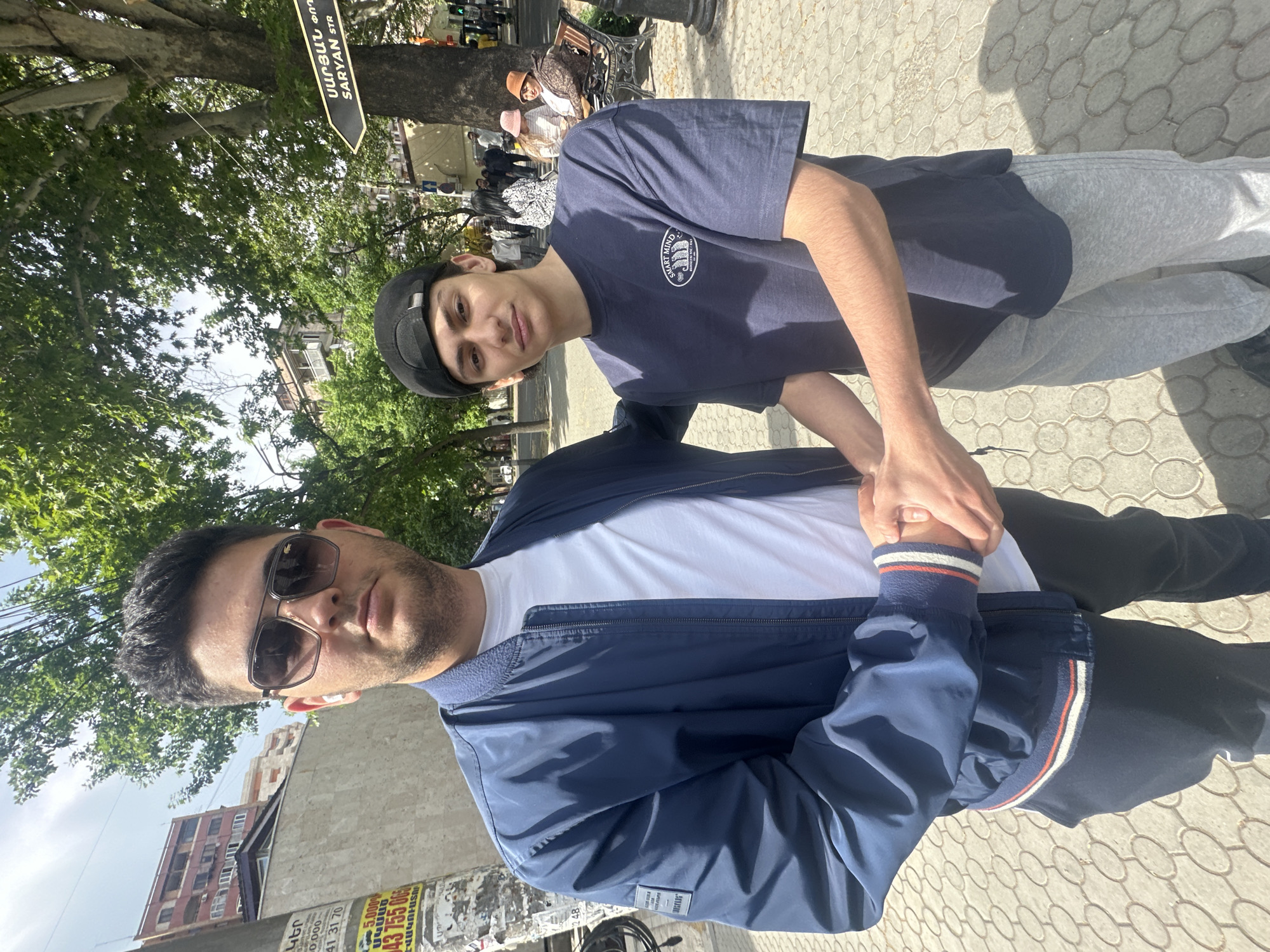 Two young men pose for a selfie on a sunny day in Yerevan, Armenia. The man in the foreground wears sunglasses, a white t-shirt, and a blue jacket with white and red stripes on the cuffs. He has dark hair and a serious expression. The man behind him wears a navy blue t-shirt, grey sweatpants, and a black cap. He has dark hair and a slight smile. Their hands are clasped together. A street sign for Saryan Street (Սարյան Փող) is visible in the background, along with trees, buildings, and other pedestrians.