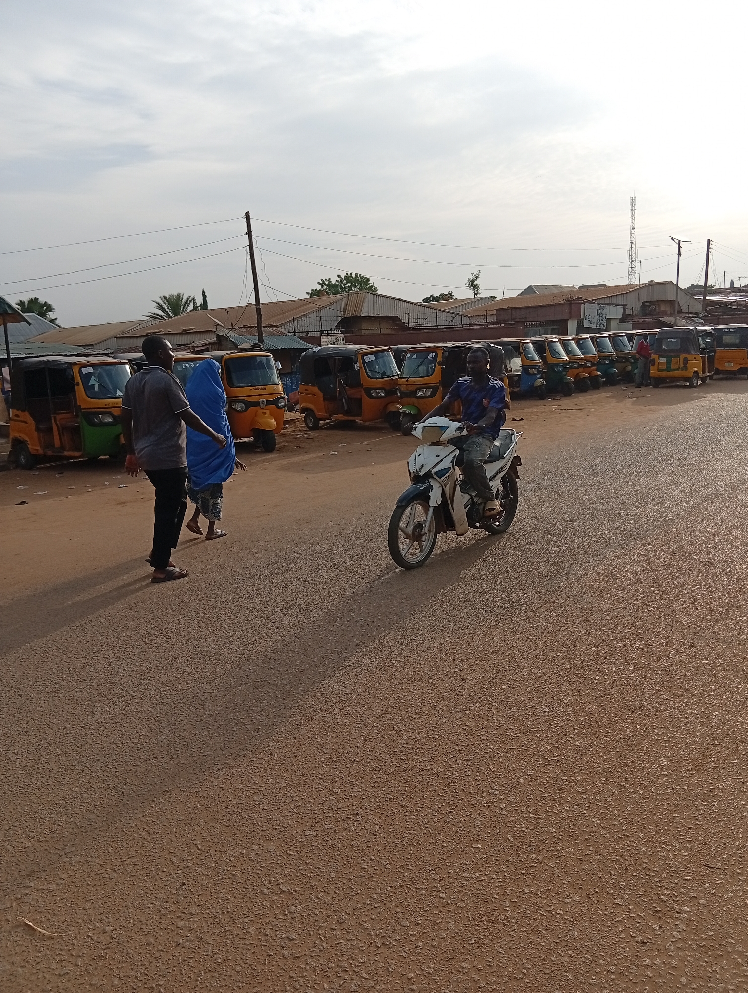 A scene unfolds on a paved road in Nassarawa, Nigeria. A row of motorized rickshaws, commonly known as "Keke Napeps," are parked on the side of the road. A man in a grey shirt and black pants walks alongside a figure cloaked in a blue hijab. A man on a white and blue motorcycle drives towards the viewer. In the background, buildings with corrugated metal roofs are visible, along with a communications tower. The sky is overcast.