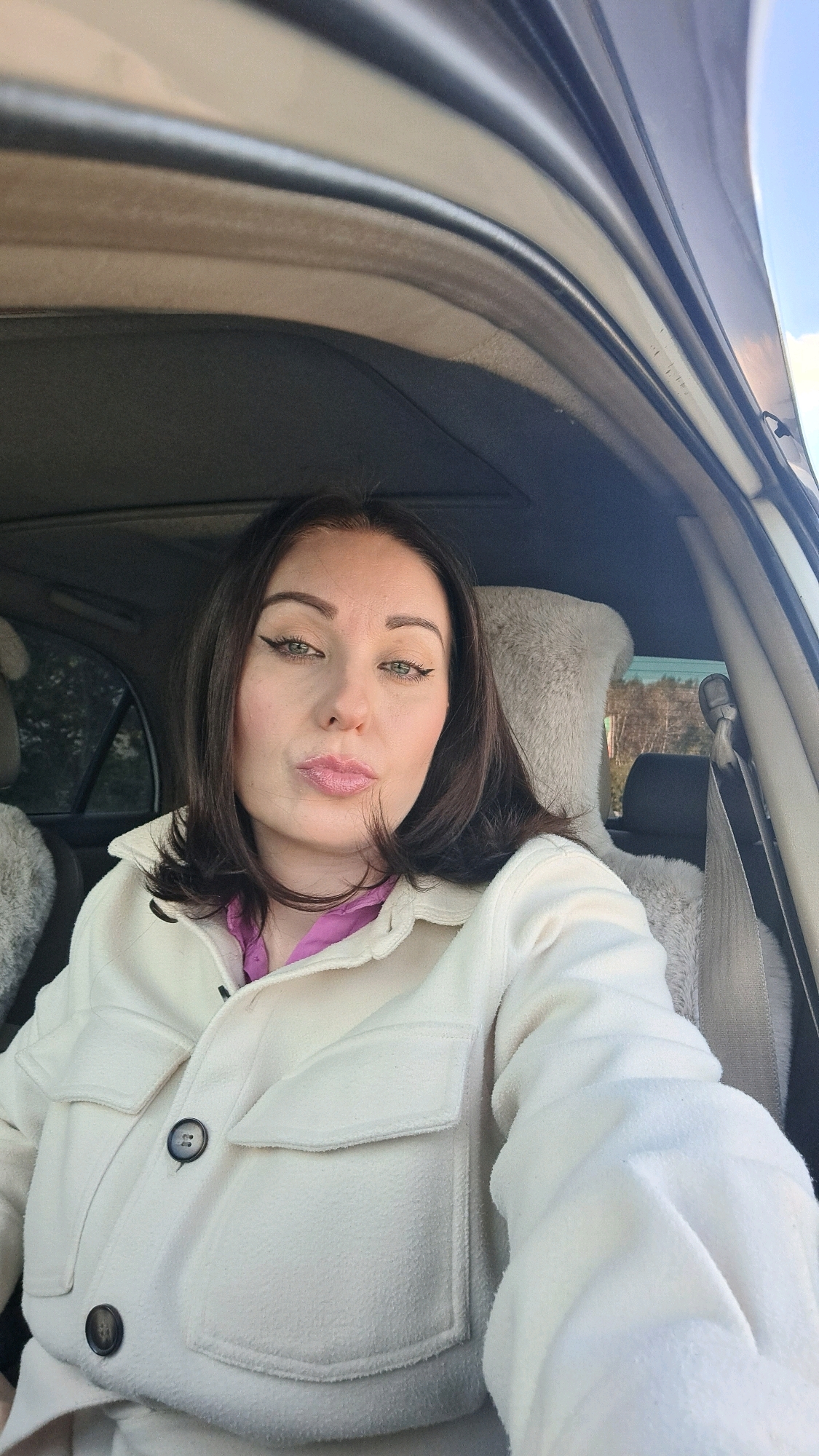 A woman with dark hair and full lips wearing a light-colored jacket over a pink shirt sits in the driver's seat of a car. Her eyes are open and looking at the camera, with winged eyeliner visible. The interior of the car is visible, including the headrest and seatbelt. The background outside the car appears to be a natural setting with trees under a blue sky. The image appears to be a selfie taken in Irkutsk, Russia.