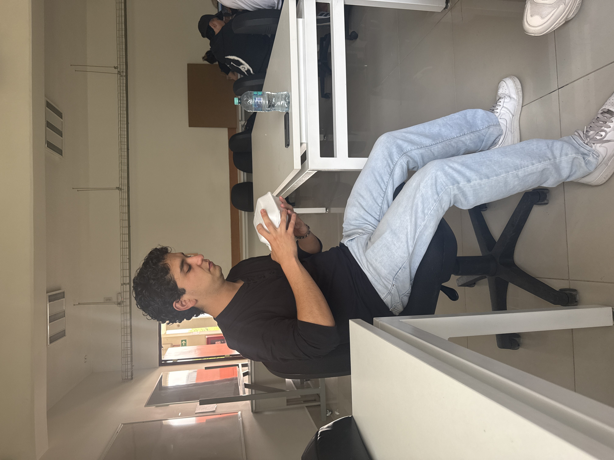 A young man with curly dark hair sits on an office chair in what appears to be a classroom or office space. He is wearing a black long-sleeved shirt and light blue jeans, and white sneakers. He holds a white container in his hands, examining it.
The location appears to be an educational setting, given the presence of rows of desks, office chairs, and a whiteboard. Other people are visible in the background sitting at desks. A window to the outside is also visible. Based on the architecture and environment, the location is likely in San Andrés Cholula, Mexico. A water bottle is visible on one of the desks.