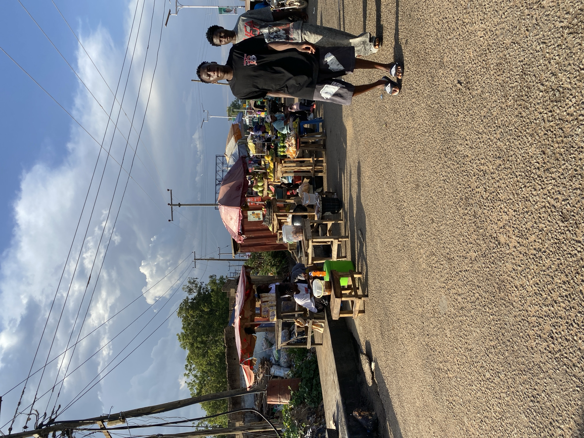 Two young men are standing on a road in Jos, Nigeria, next to a market. The man in the foreground is wearing a black t-shirt and shorts, while the man in the background is wearing a grey tracksuit. The market is comprised of several stalls selling a variety of goods, including fruits, vegetables, and household items. There are people sitting and standing at the stalls. Utility poles and wires are overhead. The sky is partly cloudy. The road is paved and appears to be a primary thoroughfare.