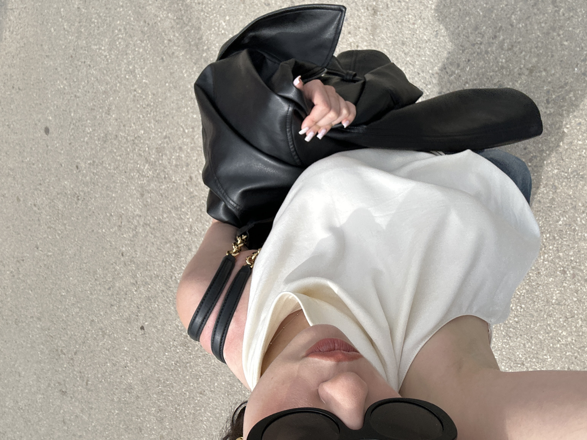 The image is a selfie taken outdoors, likely in an urban environment. A person is holding a black leather jacket in their hand, which obscures part of their body. They are wearing a white sleeveless top and sunglasses. A black handbag with gold chain details is visible. The background is a grey surface, possibly a sidewalk or pavement. No indication of the city of Sofia, Bulgaria, is evident in the image.