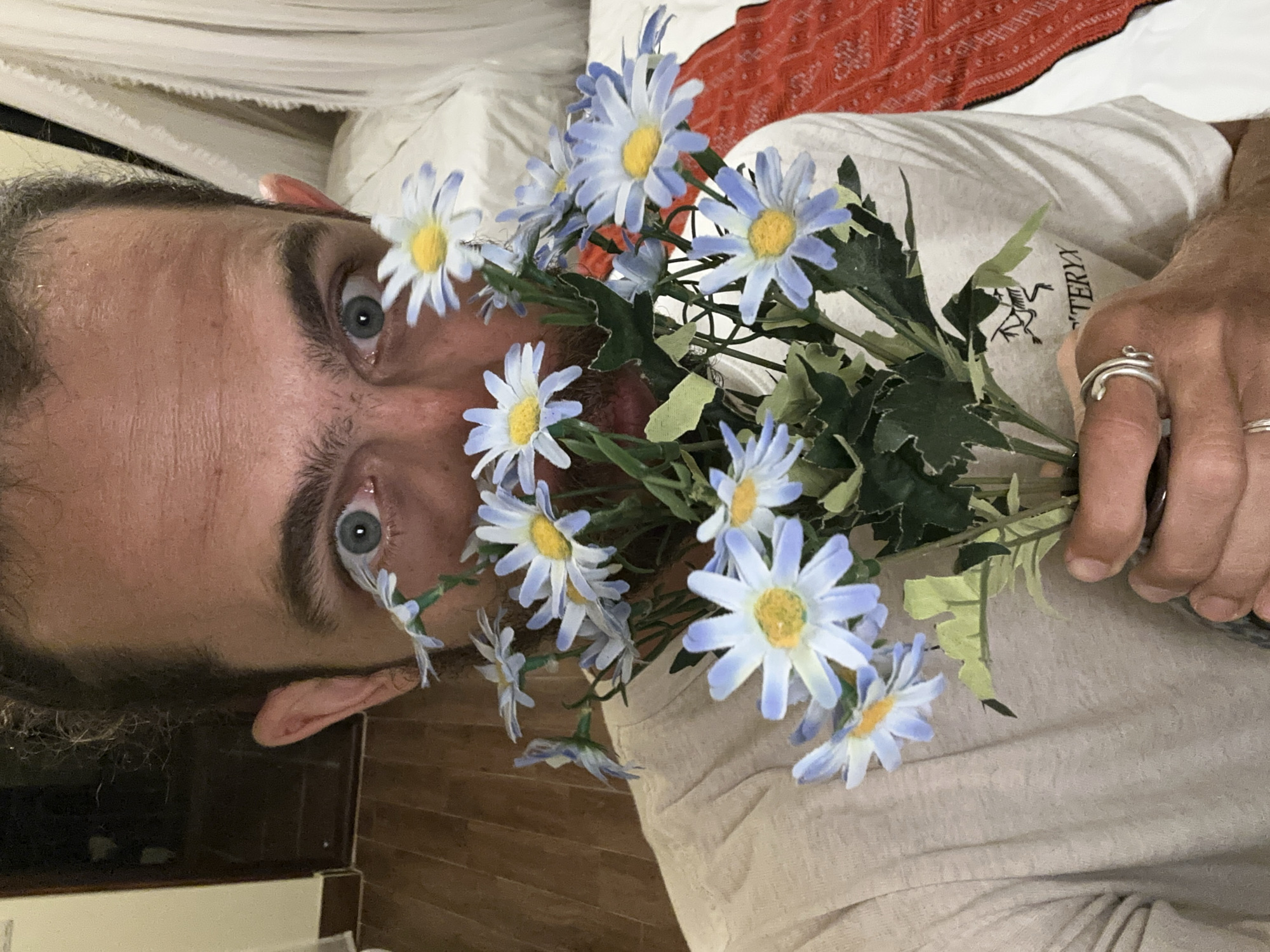 A male subject with light skin, a beard, and dark hair pulled back is positioned centrally, holding a bouquet of artificial daisy-like flowers in front of his lower face. His eyes, wide and light blue, stare directly at the viewer with a surprised or intense expression, his pupils visibly dilated. His forehead exhibits horizontal lines. He is wearing a light-colored, short-sleeved t-shirt with a black "ARC'TERYX" logo visible on the right side of his chest. On his right hand, which holds the flower stems, he wears at least two silver rings, one appearing as a double-banded design on his ring finger. The artificial flowers feature light blue petals with white tips and yellow centers, supported by green stems and leaves. The background on the left reveals a wooden floor, a light-colored wall, and a dark doorway. On the right, a bed with white bedding and a red patterned textile is visible, along with white sheer curtains or mosquito netting. The scene is indoors with artificial lighting. The image is stated to be captured in Ta Xua, Vietnam.
