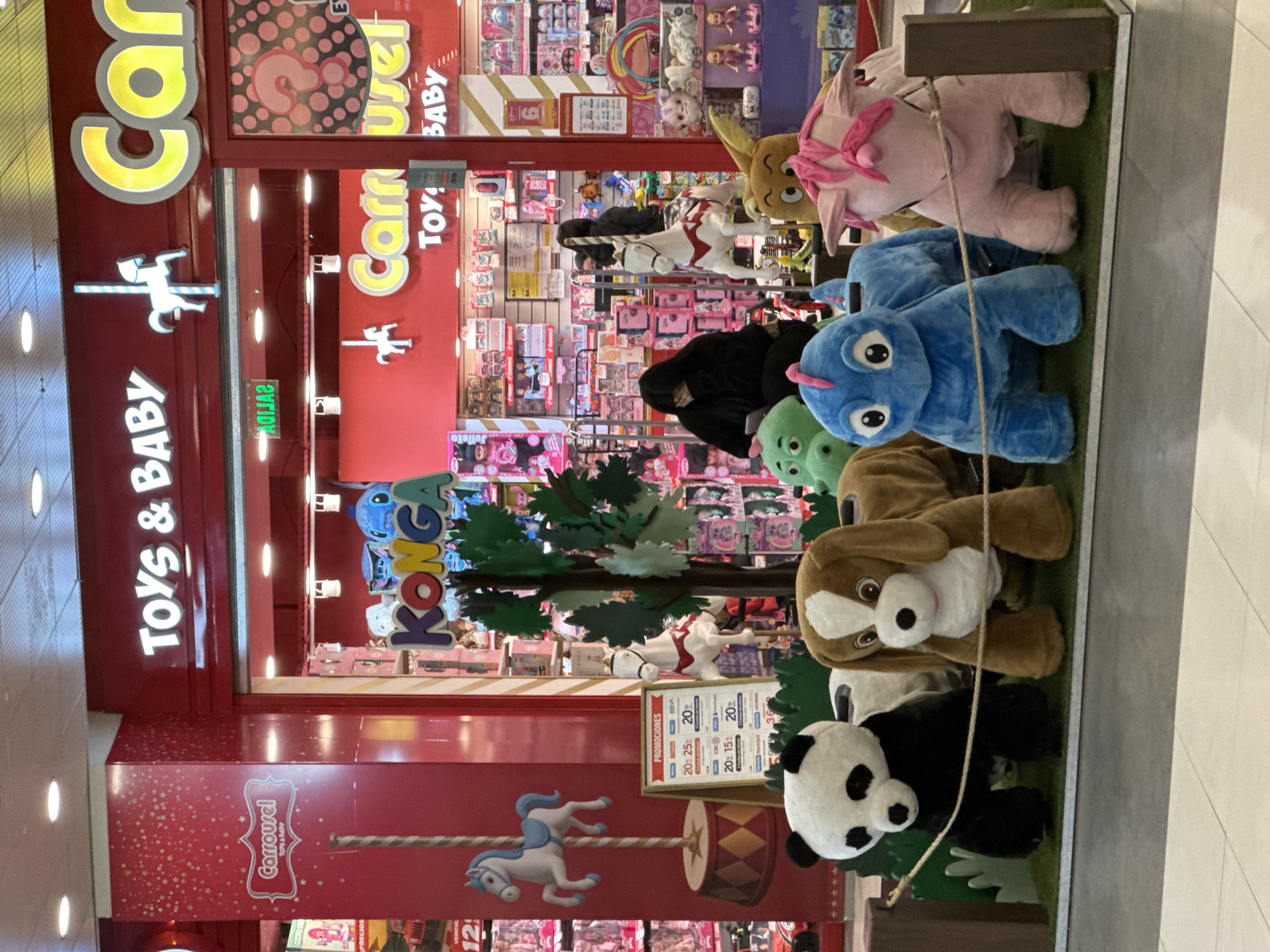 The photograph shows the entrance to the "Carrousel Toys & Baby" store, located in an indoor shopping mall. The store's signage is predominantly red and features illuminated lettering. To the right of the entrance, there's a display of stuffed animals, including a pink unicorn, a blue creature, a green creature, a brown dog, and a panda bear, arranged vertically on a patch of artificial grass within a display case. A child wearing a black headscarf is visible inside the store. In the background, shelves stocked with a variety of toys are visible. A sign promoting discounts is displayed near the bottom of the store's entrance. The location is potentially in Buenos Aires, Argentina, as indicated by the text "PRECIOS" (Prices) in Spanish and the name "Julieta," which is a Spanish name.