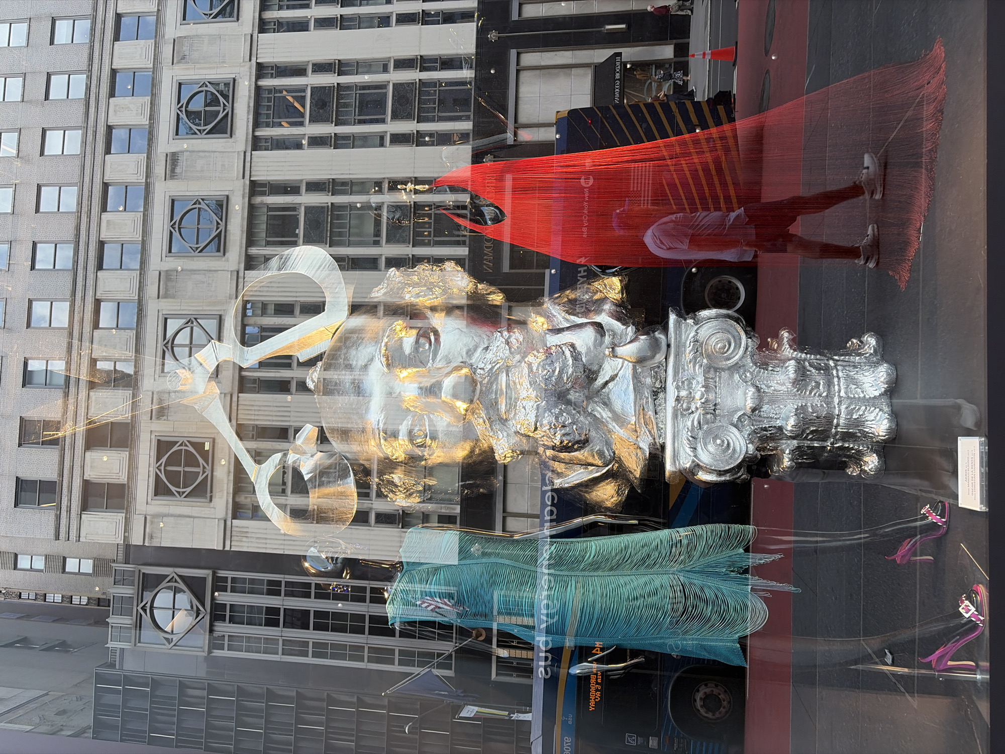This media file captures a street view through a window display, likely in a bustling urban environment. The foreground is dominated by a striking, large-scale silver sculpture that appears to be a stylized human face, possibly with classical architectural elements integrated into its design. The sculpture is partially obscured by reflections on the glass, creating a layered effect.
Behind the sculpture, a vibrant red, string-like art installation is prominently displayed. A person, likely a man, is visible within this red installation, wearing a light-colored t-shirt, dark shorts, and sneakers. He is partially silhouetted and appears to be walking or posing within the artwork. The red installation is semi-transparent, allowing glimpses of the background.
The background reveals a tall building with numerous windows, reflecting the sky and surrounding structures. Further back, a dark blue bus is visible, with some text on its side, including what appears to be "BROADWAY" in reverse. Another smaller structure with red and yellow diagonal stripes is also present.
The lighting suggests it could be late afternoon or early evening, with the sun casting long shadows and creating a golden hue on some surfaces, as seen on the silver sculpture. The scene evokes a sense of vibrant city life and artistic expression. The presence of a bus and the architectural style of the building suggest a location in a major city, consistent with the provided location context of New York, USA, specifically Broadway.