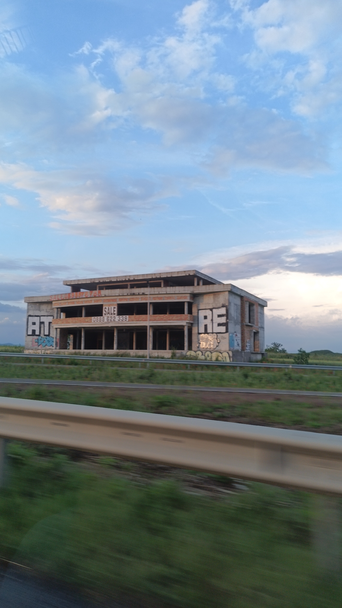 The image shows an unfinished, abandoned building with graffiti on its exterior walls, partially obscuring the windows. The words "AT", "SALE 0888 622 339", and "RE" are sprayed on the walls. The building is near a green field and a road with a guardrail, with an expansive blue sky with clouds in the background. The location is likely near Burgas, Bulgaria, based on the phone number. The scene appears to be taken from a moving vehicle.
