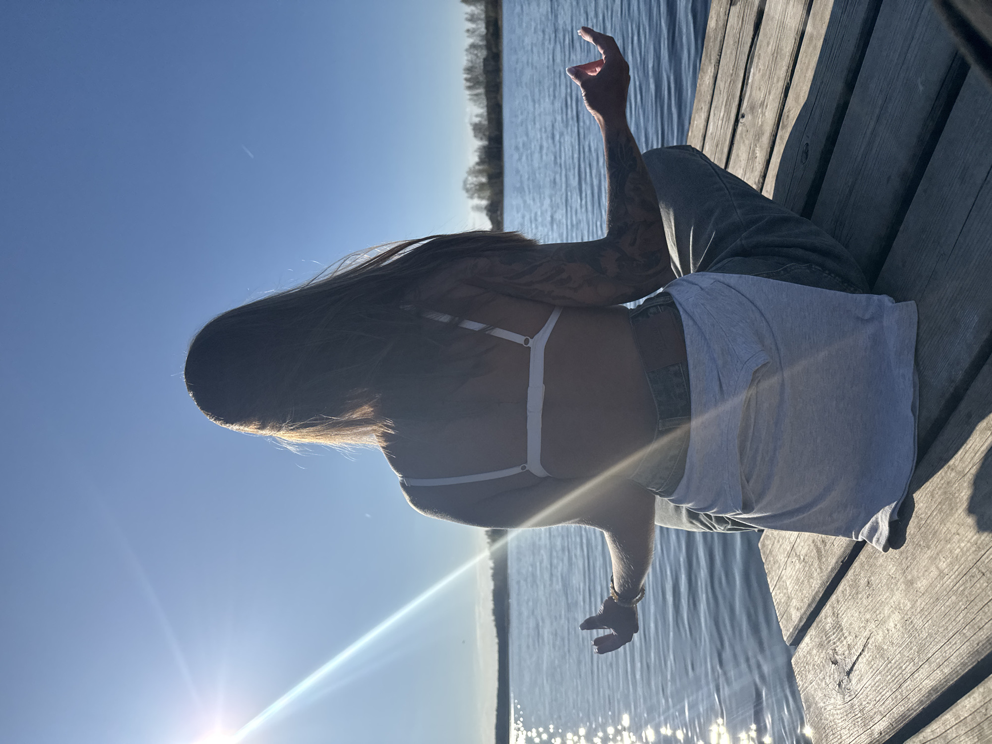 The photo shows a person sitting on a wooden dock next to a body of water, likely in Bol'shoye Kozino, Russia. The person has long hair and tattoos on their arm. They are wearing a white top and jeans, with a grey t-shirt draped over their legs. One hand is raised towards the sky. The sun is shining brightly, creating lens flare in the image and reflections on the water. The background includes trees on the far side of the water. The dock is made of wooden planks.