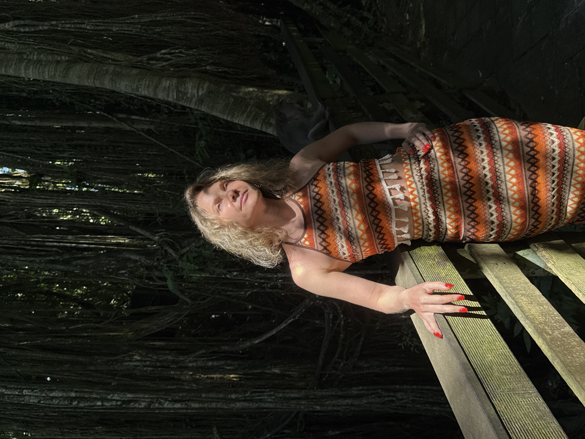 A woman with blonde, curly hair stands on a wooden walkway in a jungle environment. She is wearing a patterned halter top and skirt in shades of orange, brown, and white. Her left hand rests on a wooden railing, and her right hand is on her hip. Sunlight filters through the dense foliage, casting shadows on the scene. In the background, a monkey sits on a railing. The location is the Monkey Forest in Ubud District, Indonesia.