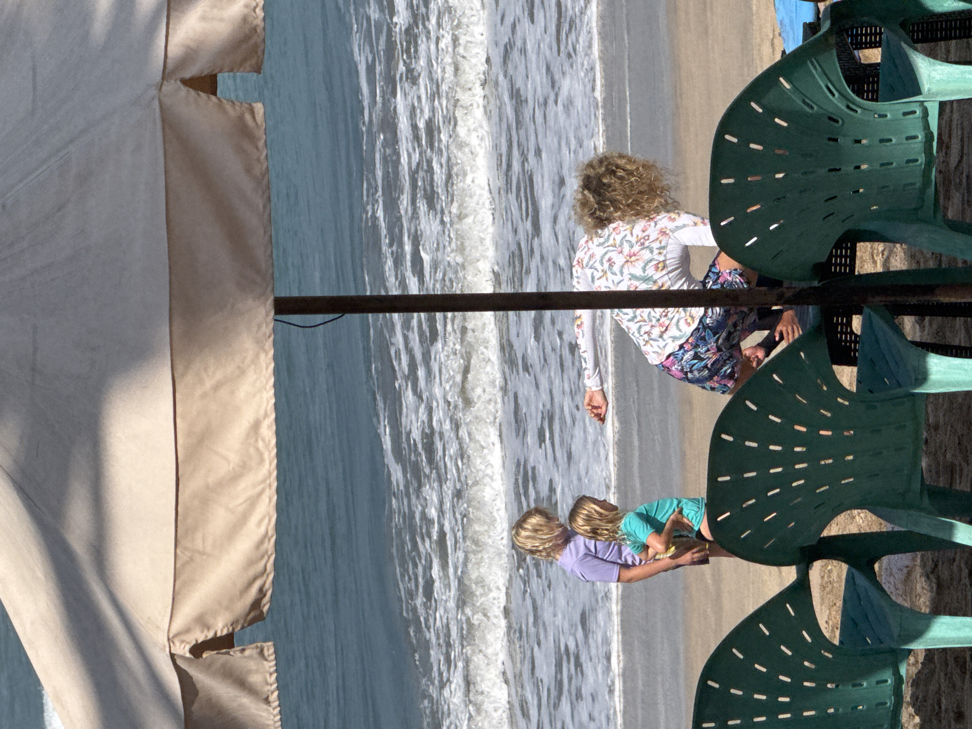 At the beach in Kuta, Indonesia, three people are present. A woman with curly blonde hair is crouched down near the water, wearing a floral-patterned long-sleeved shirt and floral shorts. In front of her, two young children with blonde hair are standing on the sand. One child wears a purple t-shirt and shorts, and the other wears a teal t-shirt and black bottoms. In the foreground, two green plastic chairs are visible. Behind the people, the ocean with moderate waves is present, and a large beige beach umbrella is overhead.