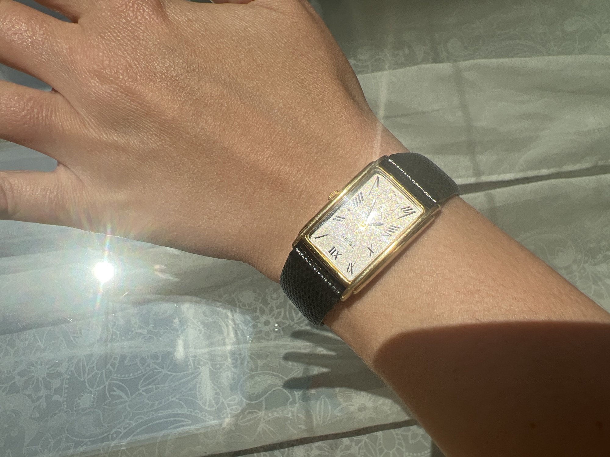 A person's left forearm and hand are visible, wearing a rectangular gold-tone watch with a black leather band. The watch face has Roman numerals and a textured, shimmering dial. The person's skin appears tanned. The background consists of a sheer white curtain with a subtle floral pattern, illuminated by bright sunlight which creates lens flare and shadows on the curtain and forearm. The scene is indoors, likely near a window. No specific location in Minsk, Belarus can be identified.