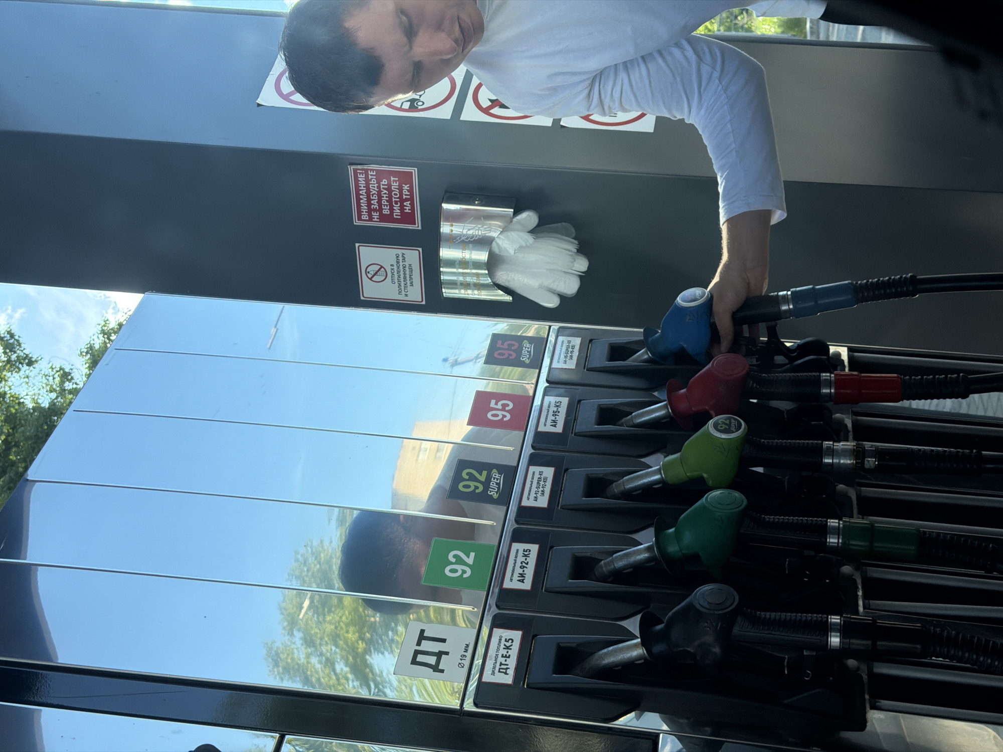 A person in a white long-sleeved shirt is holding a blue gas pump nozzle at a gas station. The gas station offers various fuel types, including ДТ (diesel), 92, 92 SUPER, 95, and 95 SUPER. Each fuel type has a corresponding colored nozzle (black, green, green, red, blue). The gas station is located in Chaikovsky, Russia. Safety signs are posted, including a "No Smoking" symbol and a reminder to return the nozzle to the dispenser. There are disposable gloves in a metal holder. The sign also says "Attention! Do not forget to return the pistol to the TRC".