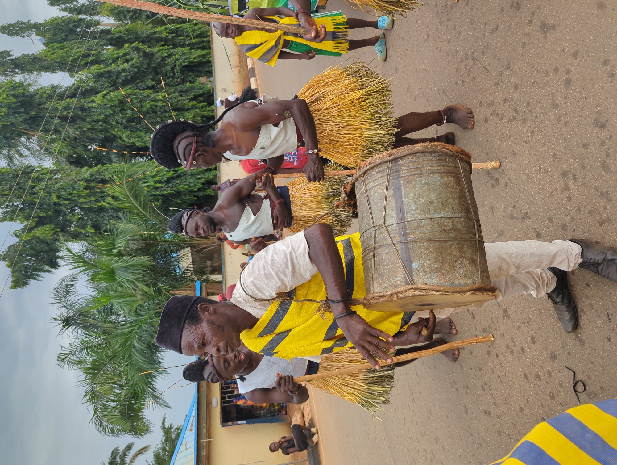 Several people are on a street in what appears to be a parade or celebration in Maigizo, Nigeria. At the forefront, a man wearing a black cap, a white shirt, a yellow and black striped garment, and off-white pants is holding a drum that he is playing. Another man behind him wears a white garment and a straw skirt. He is holding a wooden instrument and is barefoot. A third man with similar attire follows behind the second man. The setting is a paved street with trees in the background. There are buildings in the background, and the overall environment indicates a city street.