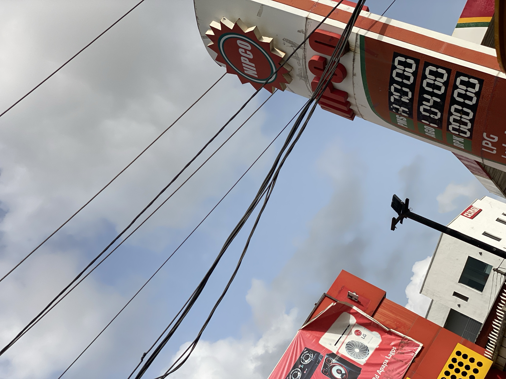 The image captures a street scene in Ado, Nigeria, looking upwards at a NIPCO gas station sign. The sign displays the price of PMS at 170.00, AGO at 104.00, and DPK at 000.00. Electrical wires crisscross the view, obscuring the sky partially. Below the gas station sign is an LG appliance advertisement poster with text indicating "Rd Apapa Lagos". In the background, there's a partial view of a white building with a sign reading "CASERTA".