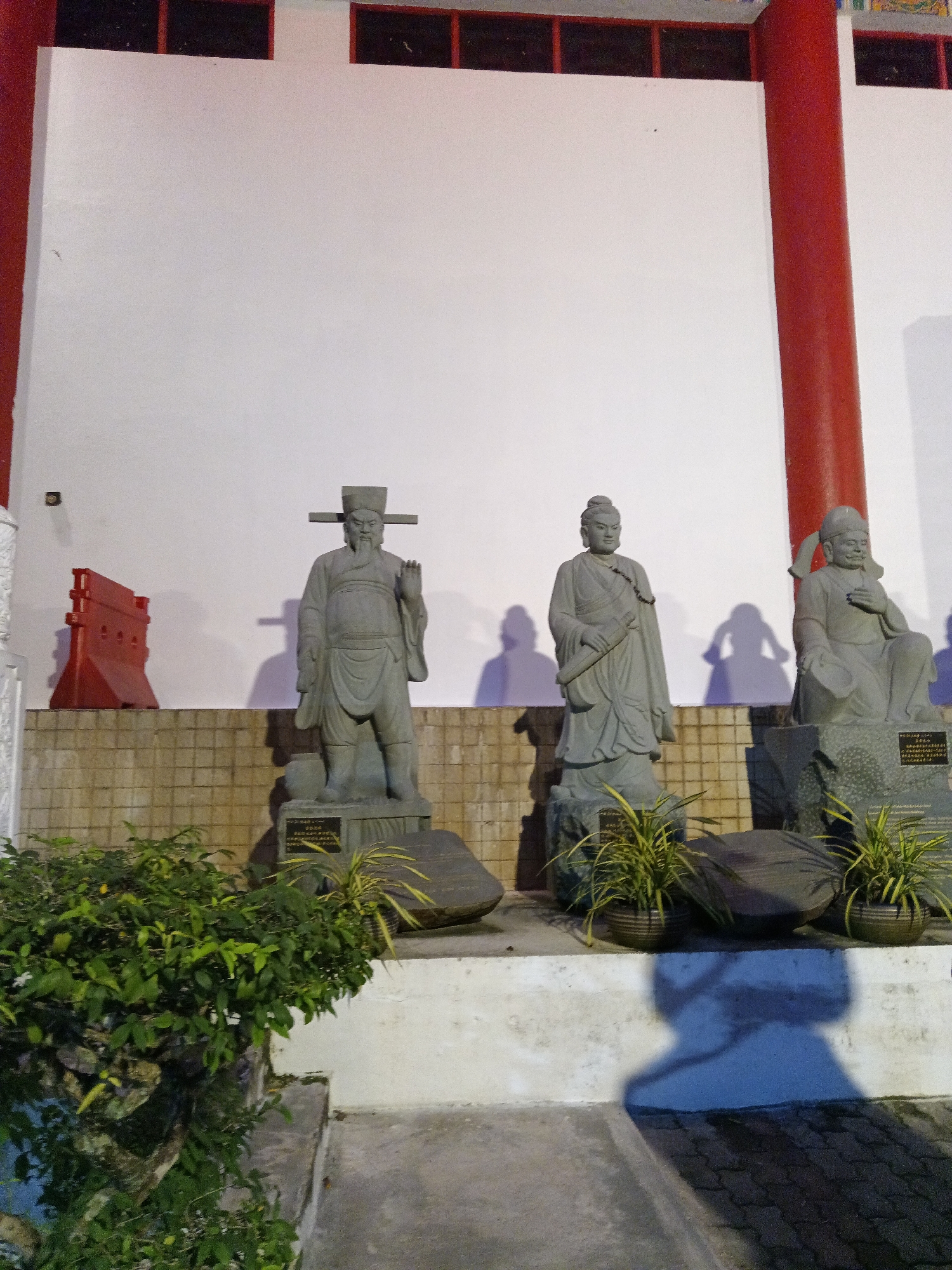 The image depicts a nighttime scene featuring several stone statues in front of a white wall, with prominent red pillars framing the right side of the scene. The statues are positioned on a tiered platform with a textured tiled wall behind them. In the foreground, lush green foliage partially obscures the base of the statues.
The leftmost statue is of a standing male figure with a beard, wearing a distinctive hat with horizontal extensions and holding one hand up in a gesture. To its right, a second standing male figure is depicted, holding a scroll-like object in one hand and wearing a more subdued headdress. The third statue, on the far right, shows a seated figure with a serene expression, hands resting in its lap. Shadows cast by the statues and other unseen objects are visible on the white wall and the ground.
A red, temporary barrier is visible to the left of the leftmost statue, behind which is a dark patch that might be an opening or another structure. Potted plants with spiky leaves are placed in front of the statues, and some of the statues are situated on large, rough-hewn rocks. The ground in the foreground is paved with interlocking bricks. The overall impression is of an outdoor shrine or a public space with religious or historical significance. The architectural elements, particularly the red pillars and the style of the statues, suggest a Southeast Asian, likely Malaysian, context. The presence of an inscription on a plaque at the base of the rightmost statue further indicates a place of commemoration or religious importance.
Based on the visual cues and typical architectural styles in Kuala Lumpur, this scene is likely located at a Buddhist or Chinese temple, such as the Thean Hou Temple, which features similar statues and architectural elements.