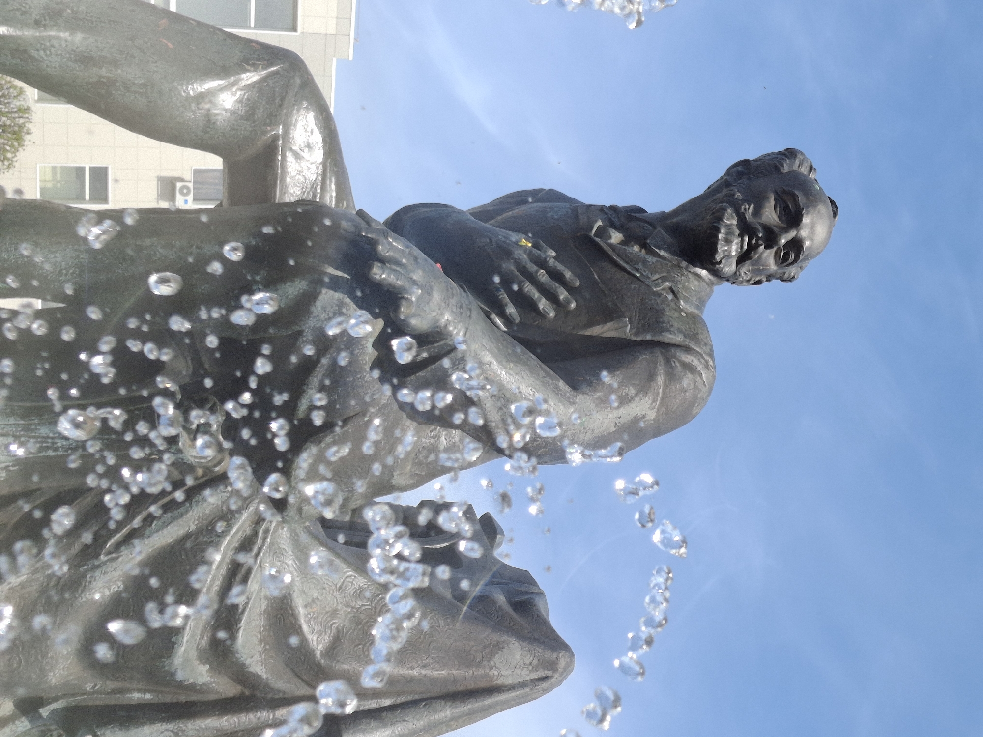 The image depicts a bronze statue of the Russian composer Pyotr Ilyich Tchaikovsky located in the city of Chaikovsky, Russia. The statue is positioned in front of a clear blue sky. A building with windows is visible in the background. In the foreground, numerous water droplets are caught in mid-air, suggesting that the statue is part of a fountain. The statue shows Tchaikovsky in a seated pose with his hands clasped in front of him. The details of his face, beard, and clothing are visible, even through the spray of water. The overall perspective of the image is from a low angle, looking upwards at the statue.