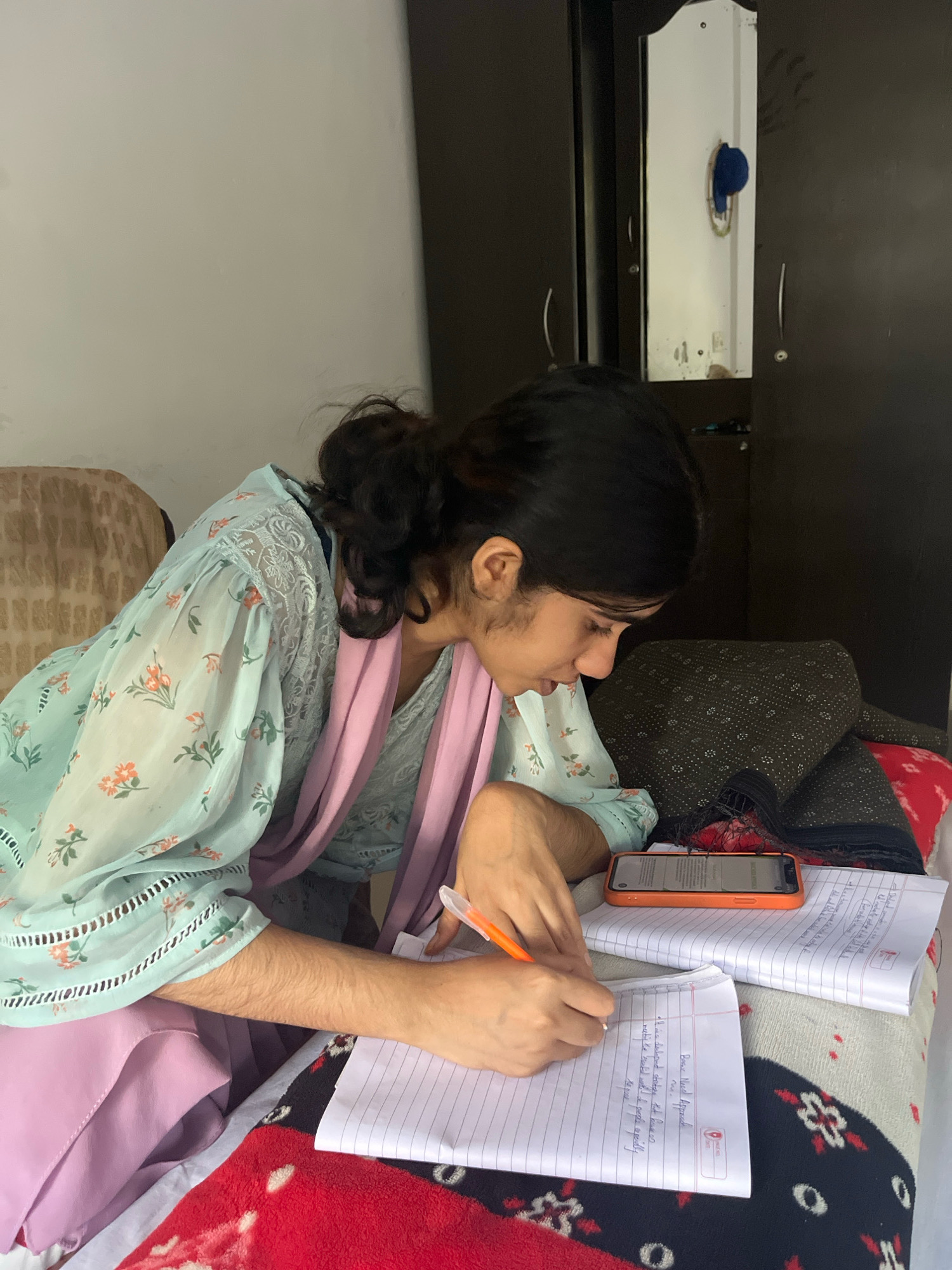 An Indian woman is seated on a bed, engrossed in writing in a notebook. She is wearing a light blue, floral-patterned top and a pink scarf. Her hair is tied back in a bun. Beside her, an orange smartphone is placed on top of some papers. The room appears to be a bedroom with a wardrobe visible in the background. The bed is covered with a red and black patterned blanket.
