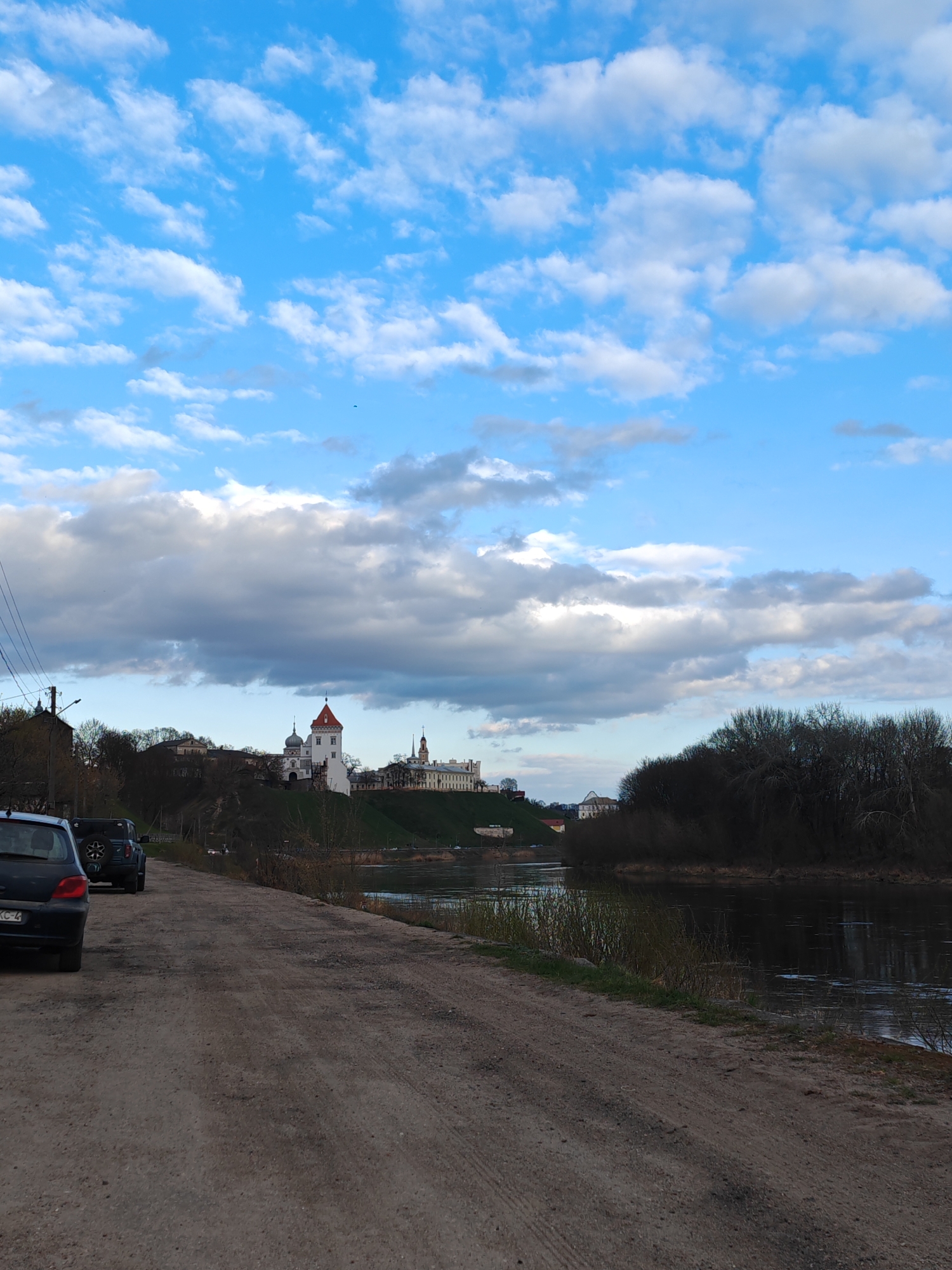 The image depicts a dirt road alongside the Neman River in Hrodna, Belarus. Two vehicles are parked on the left side of the road, a car and an SUV. On the opposite side of the river is a grassy hill, atop which stands the Old Grodno Castle with a red-roofed tower and other buildings. Trees are visible both on the hill and along the riverbank. The sky is predominantly blue with scattered white and grey clouds. Electrical wires are strung along the left side of the frame.
