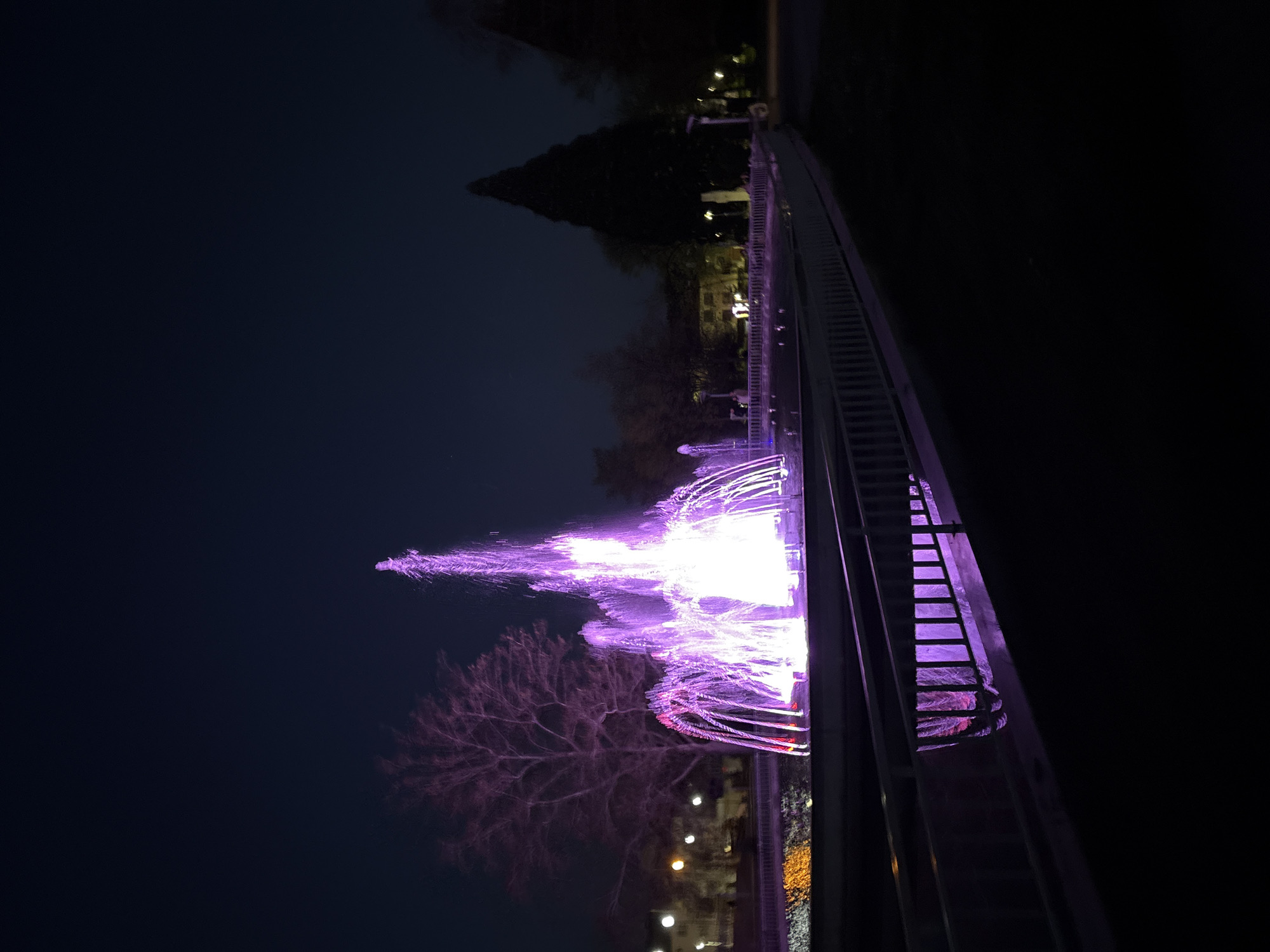 A long exposure photograph shows a fountain lit with purple lights at night in Panagyurishte, Bulgaria. The fountain is to the left of a dark metal railing. Bare branches of a tree are visible beneath the fountain's spray. Streetlights and buildings are visible in the background. The sky above the fountain is a deep blue.