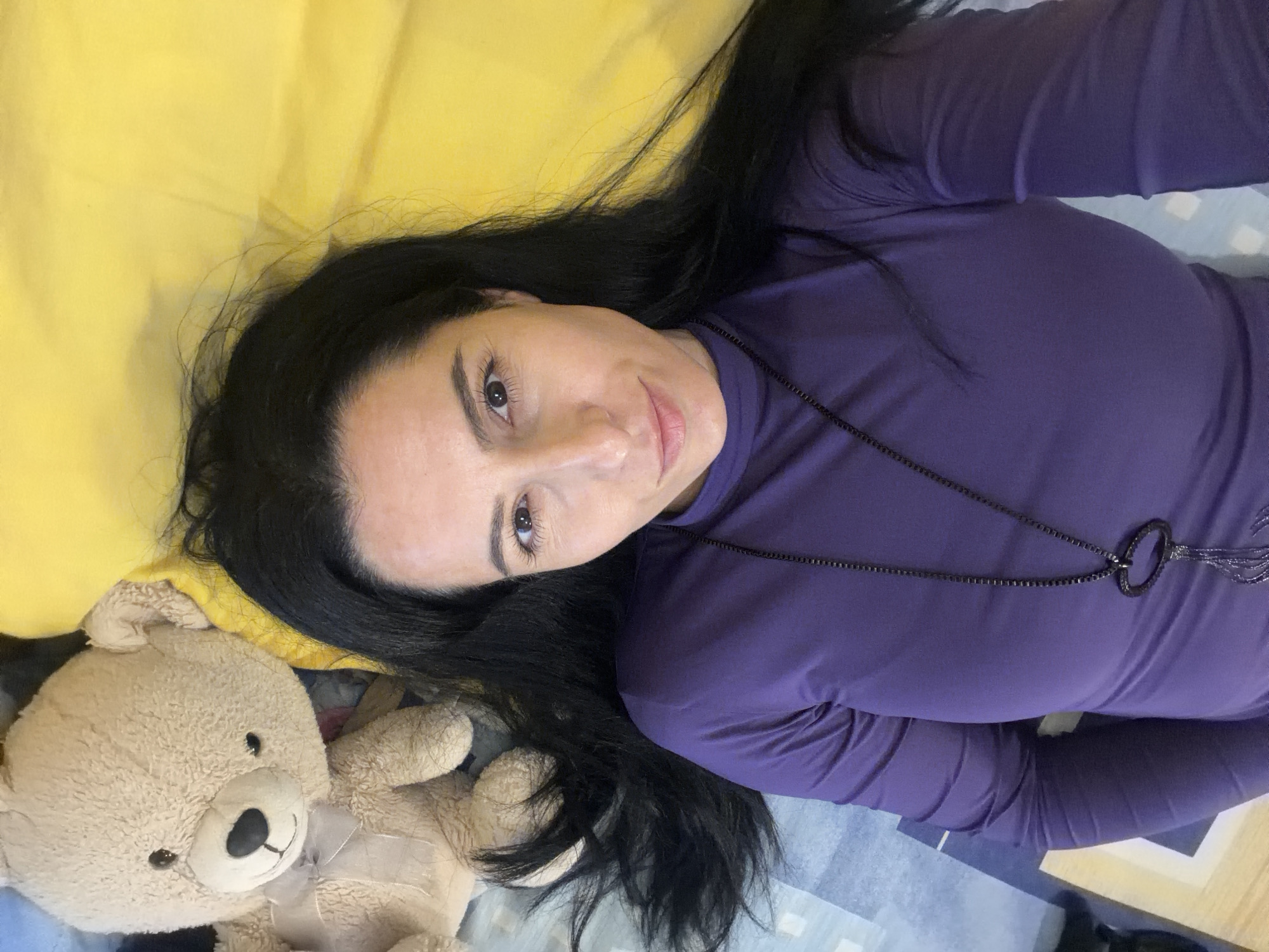 A woman is lying on a bed with a teddy bear. The woman has dark hair, light skin, and is wearing a purple long-sleeved shirt and a necklace. She is looking at the camera. To her left is a stuffed teddy bear. The background is a yellow blanket or sheet and a blue patterned sheet underneath. There is no indication of Pleven, Bulgaria.