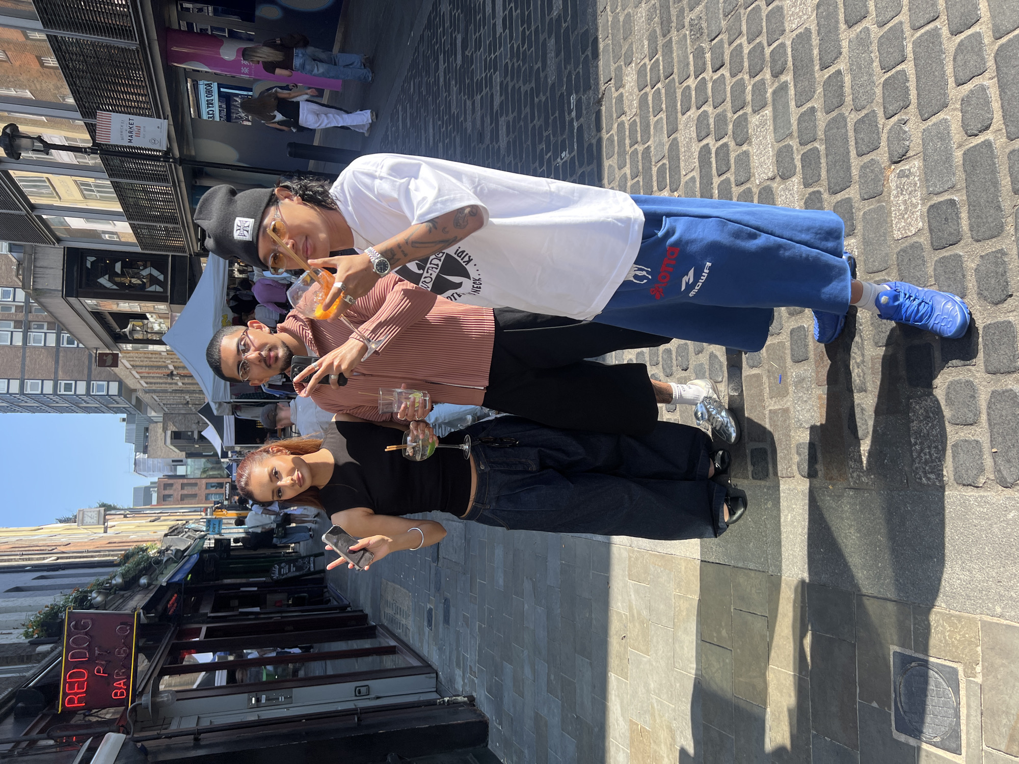 Three young adults are standing in an alley in what appears to be a sunny day in London, United Kingdom. They pose and make hand gestures for a photo, each holding a drink. The alley is paved with cobblestones, and lined with restaurants and shops. The “RED DOG” BBQ sign is prominent on the left side of the frame.