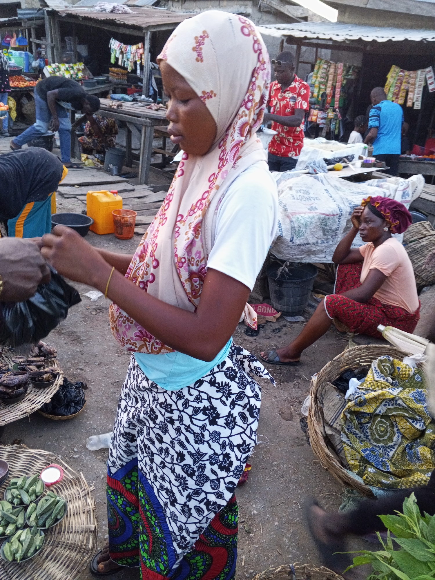 The image depicts an outdoor scene in Zango-ndaji, Nigeria, featuring a marketplace setting. In the foreground, a woman wearing a light-colored hijab, a white t-shirt, and a patterned wrap is interacting with a vendor, possibly purchasing goods being placed in a black plastic bag. Near her, there are baskets filled with items like okra and dried goods. In the middle ground, another woman is seated on the ground with a head covering and a peach-colored t-shirt. Behind her are sacks and other market items. In the background, other individuals are visible, including a man in a red patterned shirt and others near market stalls. The environment appears to be a bustling market area with temporary structures and various items for sale.