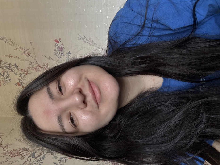 A female individual is depicted in a chest-up portrait, positioned indoors in the city of Kurovskoye, Russia. She has long, dark, straight hair, parted centrally, framing her face and falling over her shoulders. Her face is oriented slightly to her left, looking towards the camera with a gentle, closed-mouth smile. Her skin appears to have a natural sheen, and a singular reddish mark is present on her forehead above the nasal bridge. She is wearing a blue, loose-fitting garment, possibly a t-shirt, with a faint, partially obscured light-colored inscription visible on its front. The background consists of a light-colored wall covered with wallpaper featuring a repeating pattern of dark floral branches, red flowers, green leaves, and small dark birds.