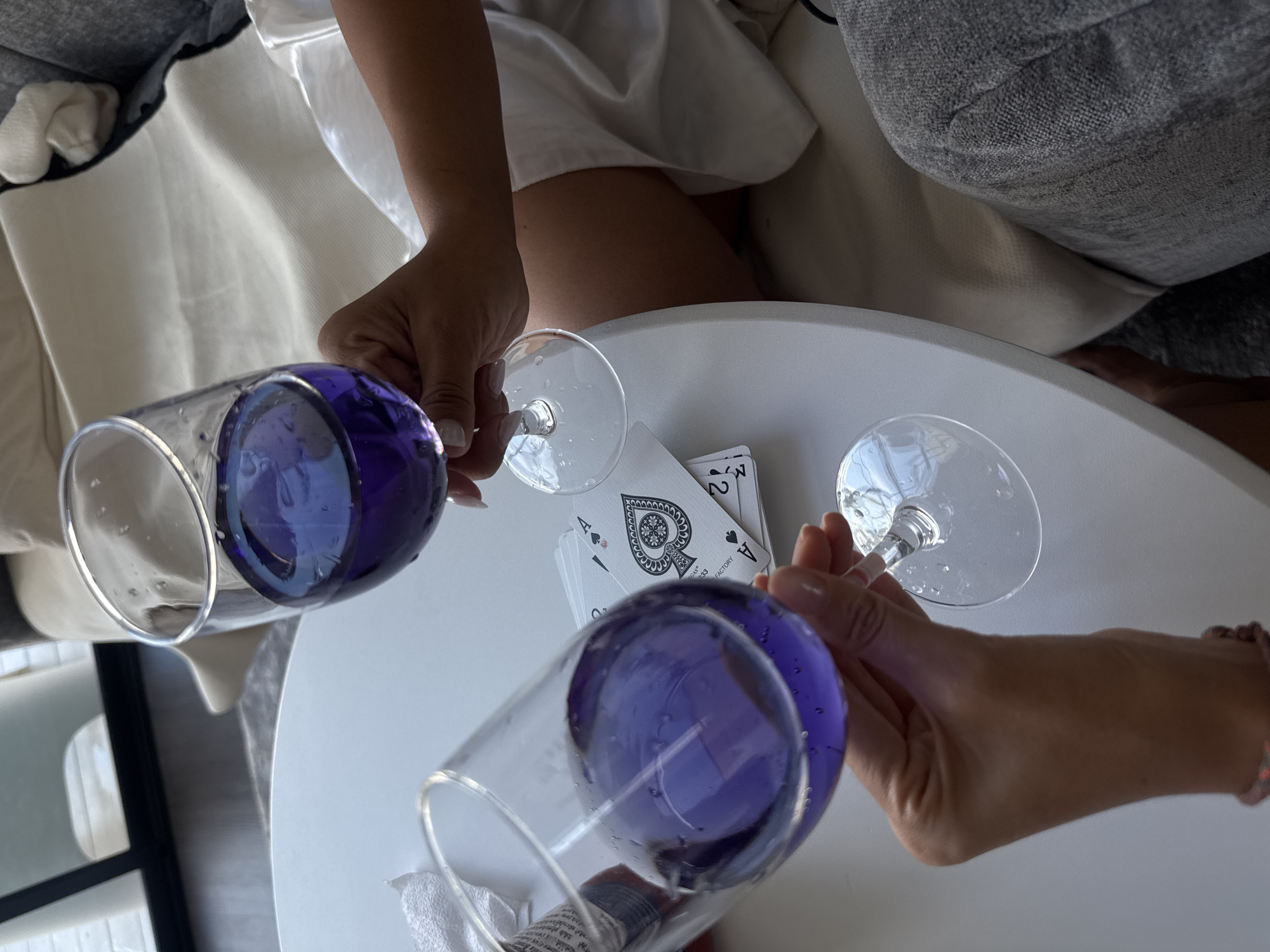 In Baan Tai, Thailand, two glasses of purple liquid are held by unseen hands over a white table. A deck of playing cards, including two aces of spades and a two of spades, lies on the table. The scene appears to be indoors, possibly in a living room, with soft seating and a reflective surface visible in the background.