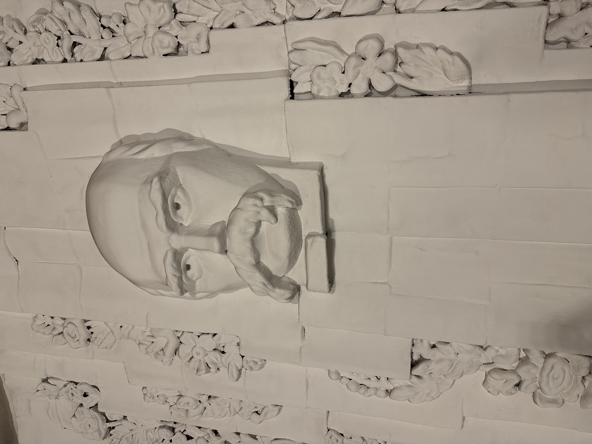The image shows a close-up of a white bas-relief sculpture of a man's head, with a prominent mustache and balding head, set against a textured background of what appear to be stacked rectangular panels with floral and leaf motifs. The sculpture is centrally located and appears to be part of a larger architectural or decorative element. The lighting casts shadows that accentuate the three-dimensionality of the relief. This sculpture is of Taras Shevchenko, the renowned Ukrainian poet and artist, and is located on the facade of the National Opera of Ukraine named after Taras Shevchenko, in Kyiv, Ukraine.