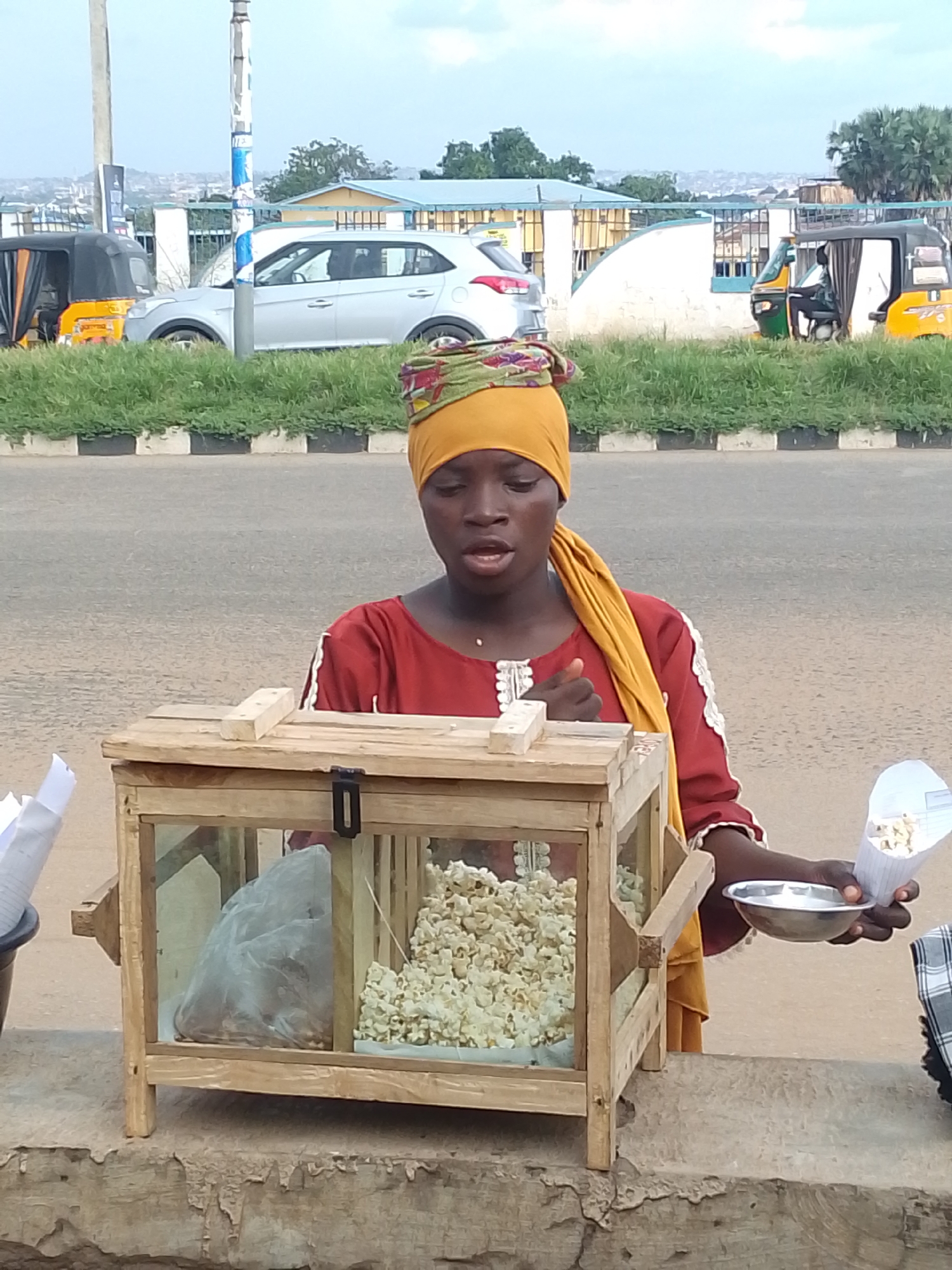 A young Black woman, wearing a red tunic with white trim and a yellow and green headscarf, stands behind a wooden and glass popcorn stand on a concrete ledge in Oja Oba, Oko Erin, Nigeria. She is holding a metal bowl in her right hand and a paper cone filled with popcorn in her left hand. The popcorn stand contains a large bag of unpopped kernels and a display of popped popcorn. In the background, a silver car and two auto-rickshaws are visible on a paved road. A grassy median with a black and white curb separates the road from the popcorn stand. Buildings and a distant cityscape are visible on a hill behind the road. The sky is overcast.