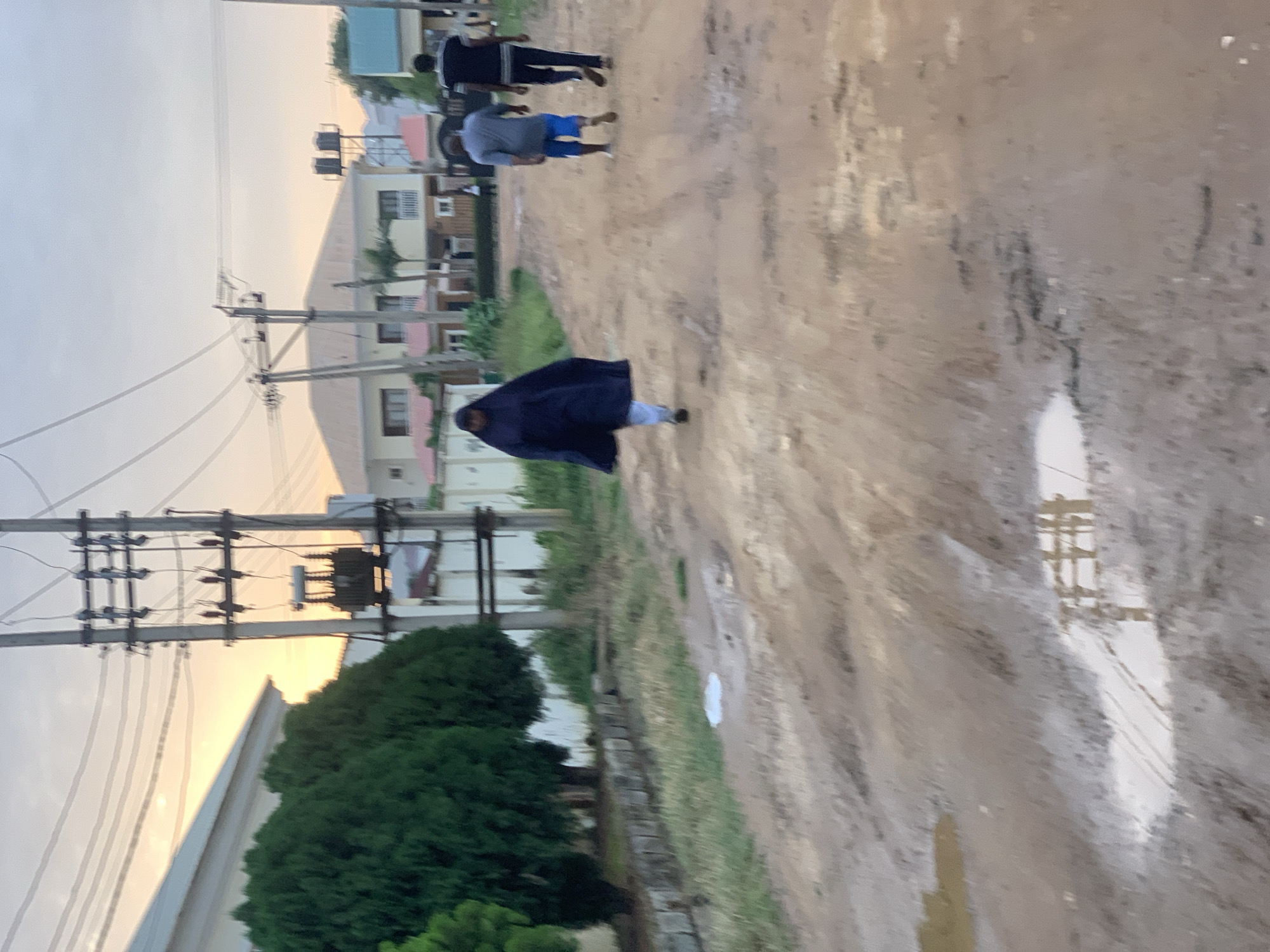 The image depicts an outdoor scene on a dirt road with several puddles of water, suggesting recent rain or inadequate drainage. The road is uneven and muddy, lined with dry grass and some greenery on the left. In the background, there are several modern-looking houses and buildings with multi-story structures. Electrical poles with wires and a transformer are visible, indicating the presence of utility infrastructure.
Three individuals are walking on the road. In the foreground, a person wearing a dark blue hijab and light-colored pants walks away from the camera. Behind them, a person in a grey shirt and blue shorts walks in the same direction. Further behind, another person in a black shirt and dark pants also walks away from the camera. The overall lighting suggests it is either early morning or late afternoon, with the sky exhibiting a soft, pale hue. The scene appears to be in a suburban area of Abuja, Nigeria, characterized by its developing infrastructure and residential architecture.