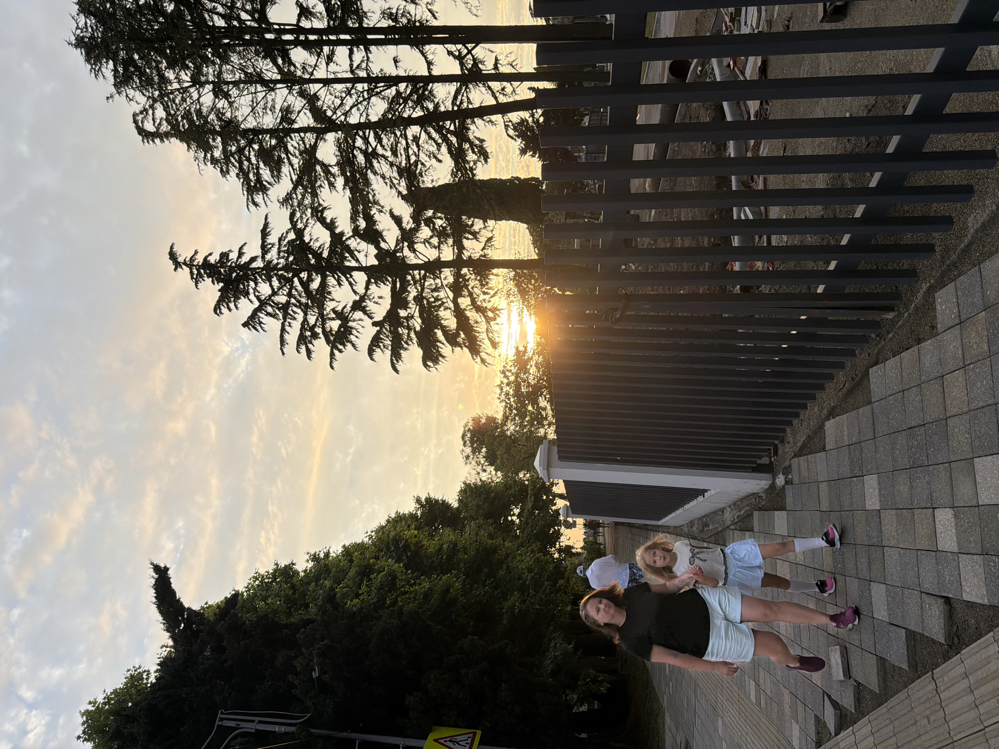 In Sochi, Russia, a woman and a young girl walk along a paved sidewalk at sunset. The woman, wearing a dark t-shirt and light-colored shorts, walks on the left side of the path, holding her left arm down. The girl, in a light-colored top and a light blue skirt with white socks, walks beside her, holding the woman's hand with her right hand. Behind them, other pedestrians are visible, including a man in a white shirt and patterned shorts walking away from the camera.
The sidewalk is constructed with square gray pavers, some of which appear to be unevenly laid or under construction. To the right of the sidewalk, a dark gray metal fence runs parallel to the path, with vertical bars and a horizontal top railing. Beyond the fence, construction materials and debris are visible, suggesting ongoing work.
The background features lush green trees, including tall evergreen trees that silhouette against the sky. The sky itself is a canvas of sunset colors, with a mix of pale yellow, orange, and pink hues from the setting sun, partially obscured by clouds and trees. The overall lighting suggests it is either early morning or late evening. A yellow triangular sign with a pedestrian symbol is visible on a pole to the far left of the image.