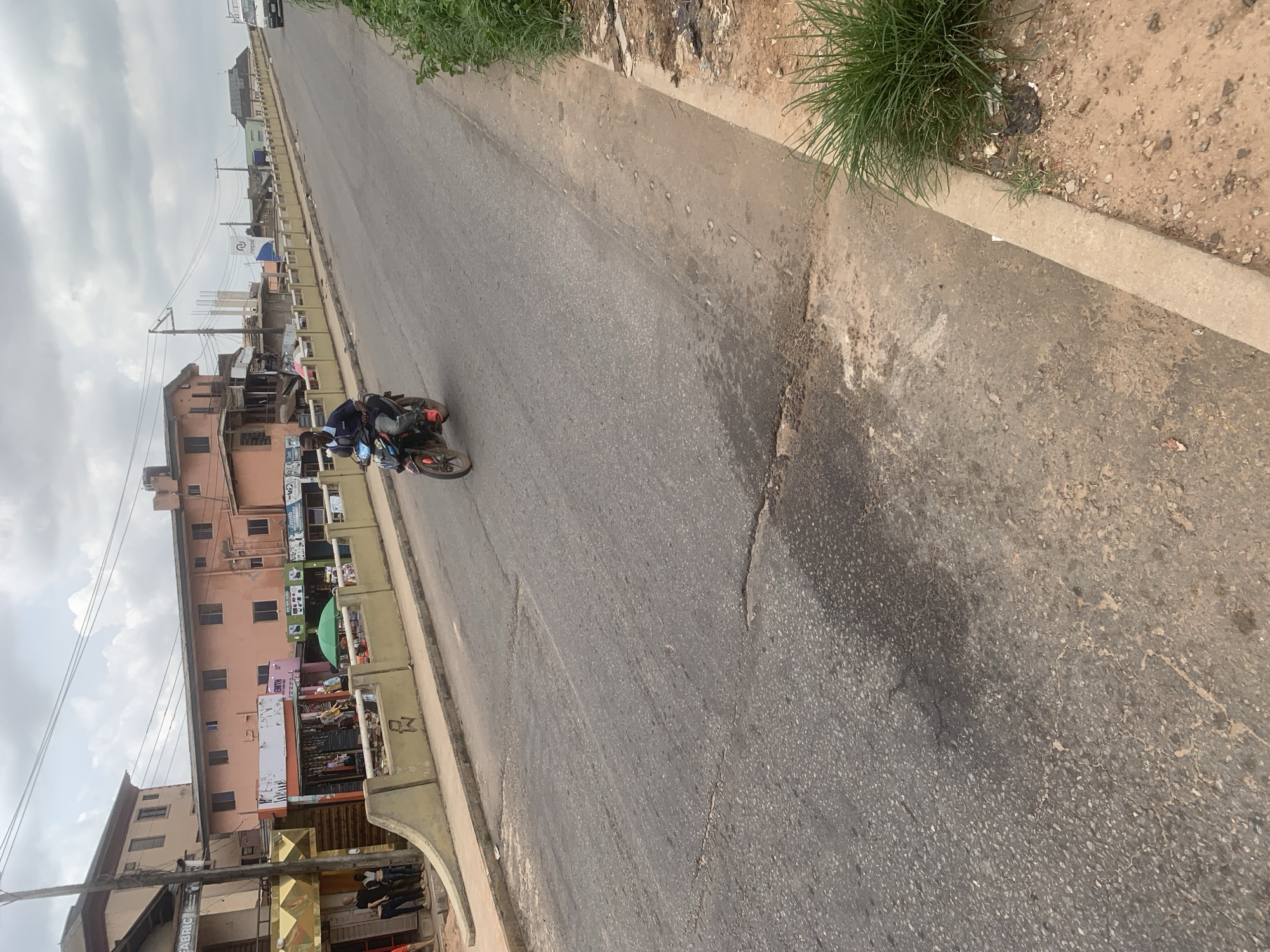 A man rides a motorcycle on a paved road in Shagamu, Nigeria. The road has several cracks and is lined with a concrete barrier. Buildings with shops are visible on the left side of the road, and lush green vegetation grows on the right. The sky is cloudy.