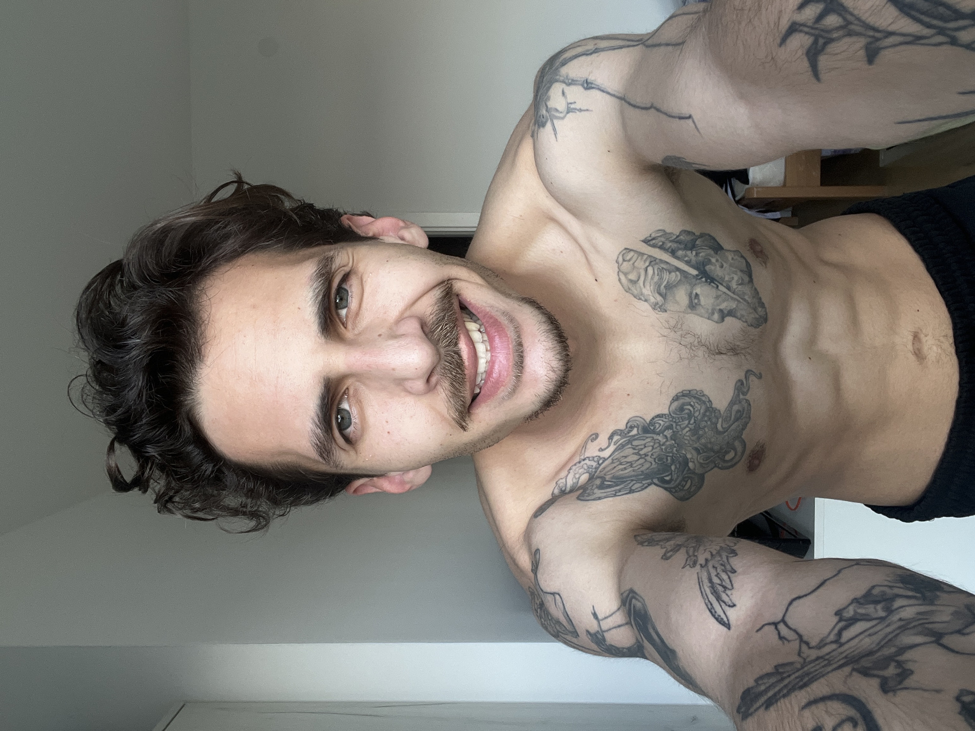 A man with dark, curly hair and a mustache smiles at the camera. He is shirtless and has multiple tattoos on his chest and arms. He is wearing black shorts. The background appears to be an interior space with a white wall and door. The photograph does not contain information to determine the location in Rafailovići, Montenegro.
