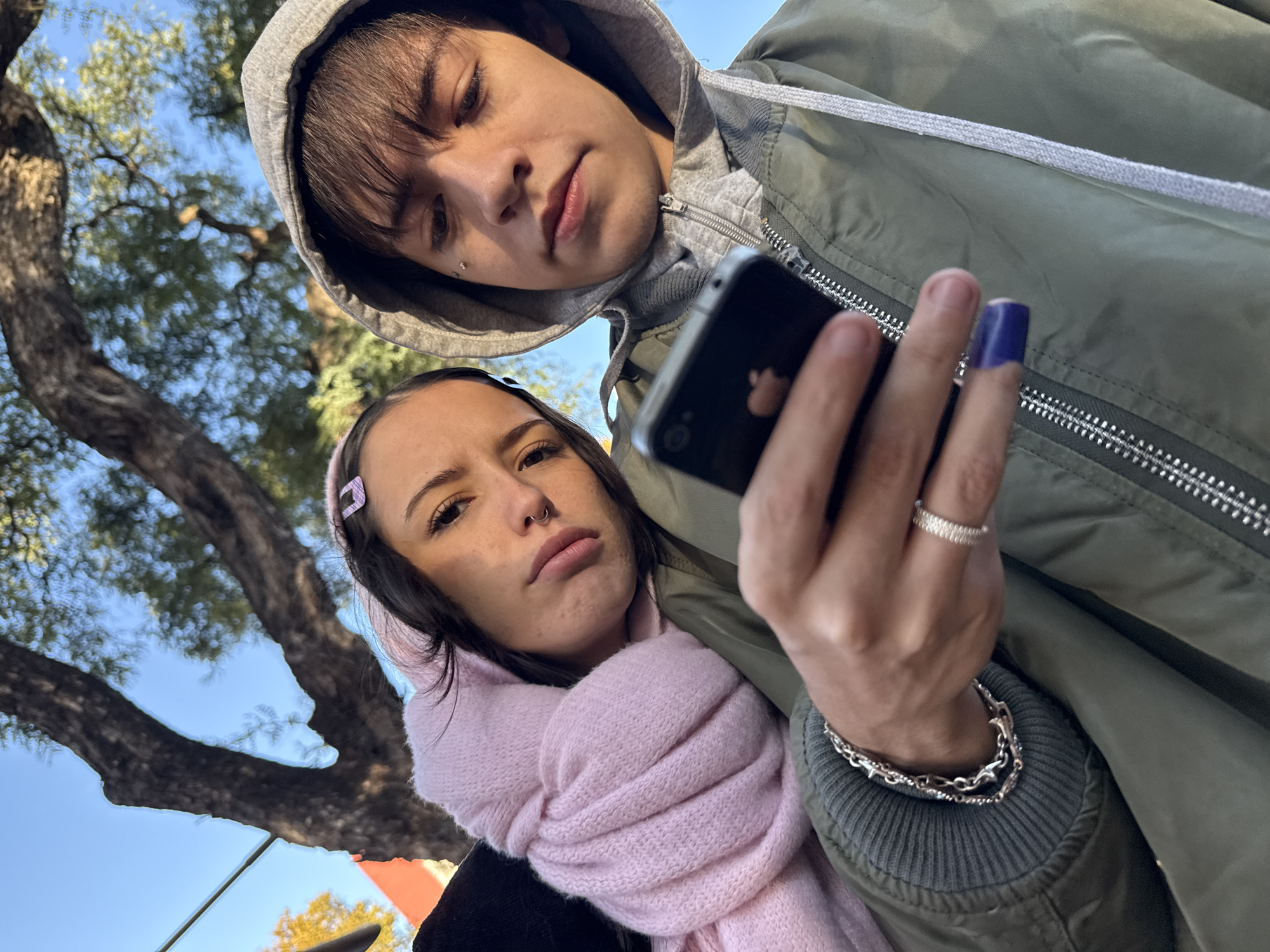The image shows two people taking a selfie outdoors. One individual is wearing a green jacket with a gray hoodie underneath, while the other is wearing a pink scarf and hair clip. A tree is visible in the background. The photo appears to be taken in the city of Buenos Aires, Argentina.