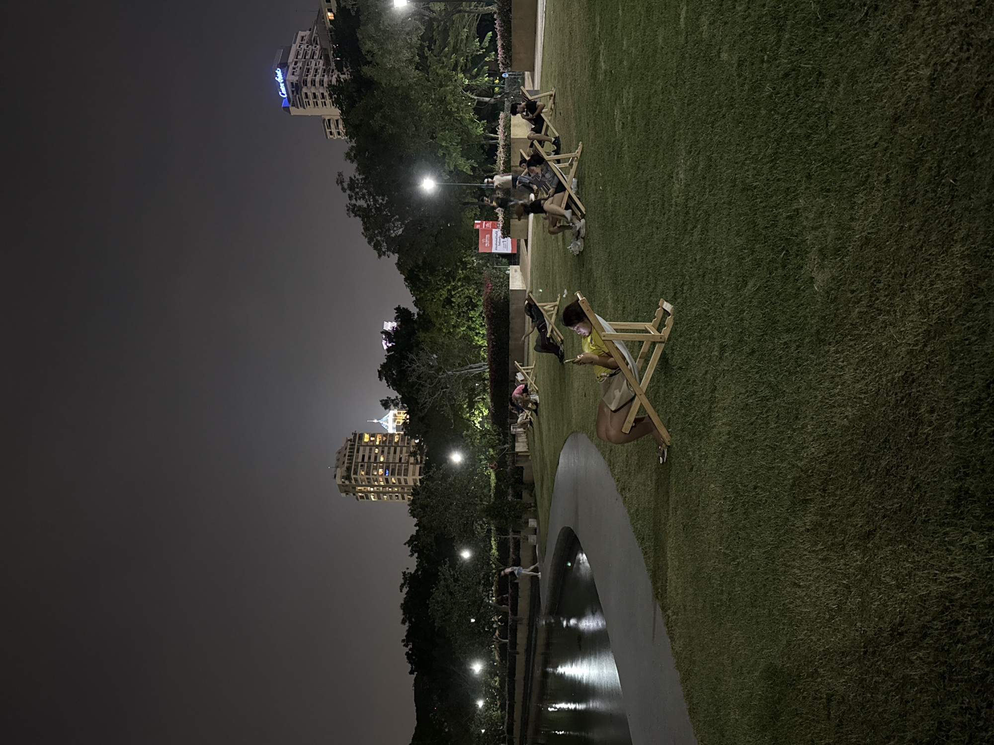 At night, a park in Bangkok, Thailand features people relaxing in folding chairs on a grassy lawn, bordered by a reflective pool and trees illuminated by streetlights. In the background, illuminated buildings rise against the dark sky. People are sitting in beach-style chairs on the right side, and on the left side is a lake or pond with trees and streetlights.
