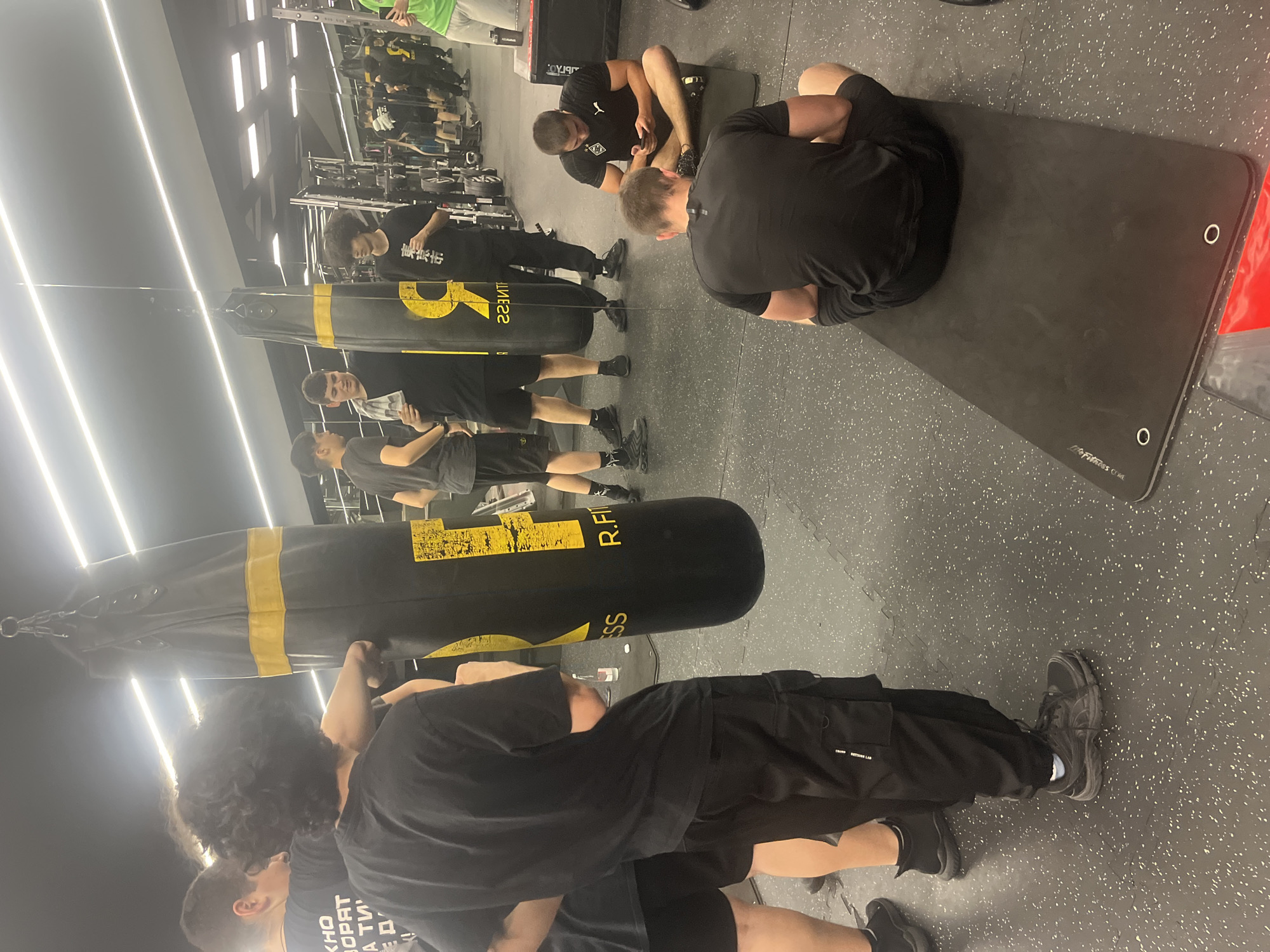 The image depicts an indoor scene, likely a gym, with multiple people engaged in various activities. Several individuals are positioned around a punching bag, possibly preparing for or engaging in boxing exercises. Adjacent to them, other people are seen performing what appear to be abdominal exercises on a mat. The gym environment is suggested by the presence of exercise equipment in the background. No specific visual indicators suggest that this location is in the city of Varna, Bulgaria.
