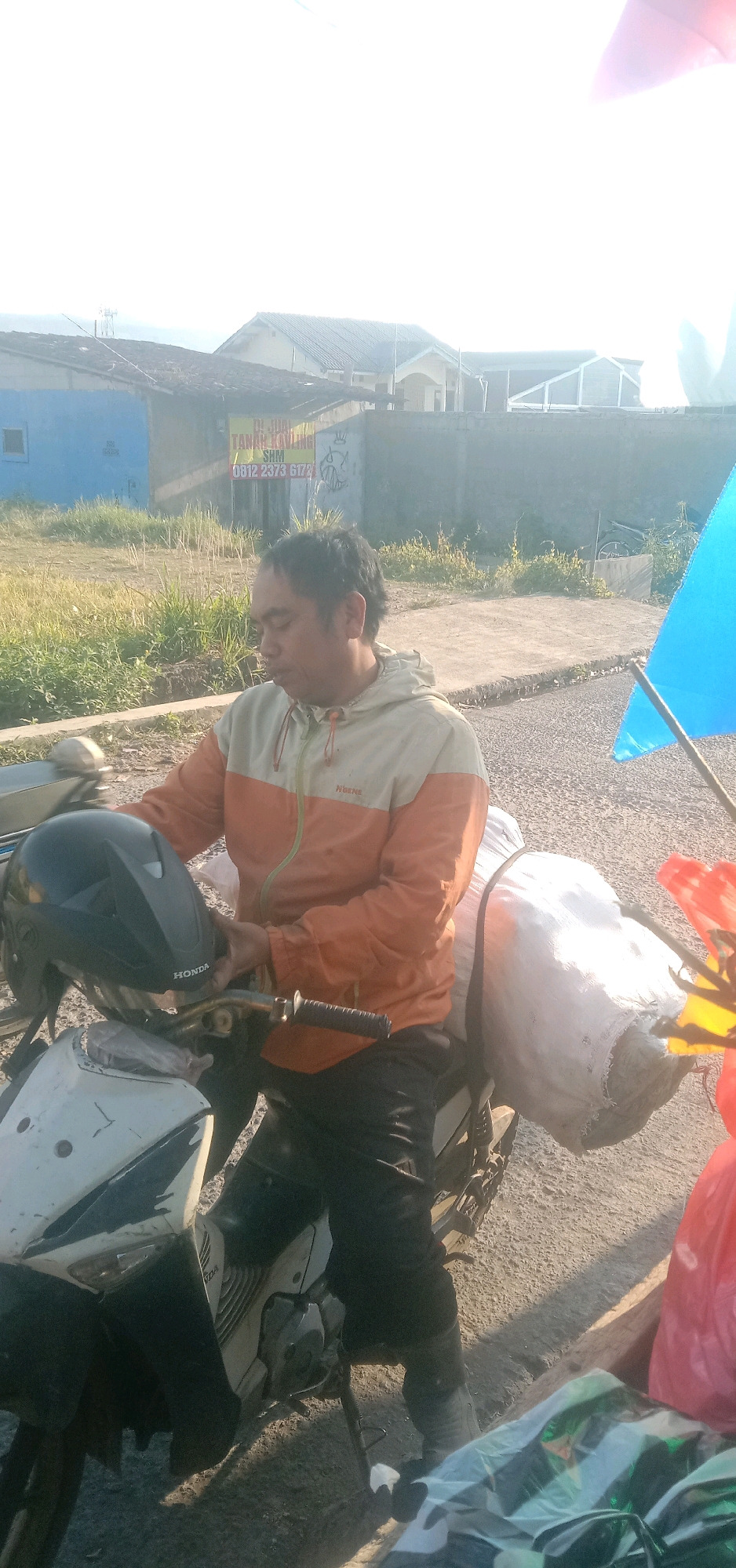 In Sukanagalih, Indonesia, a man is seated on a motorcycle. He is wearing an orange and light-colored hooded jacket and dark pants. He is holding a black helmet with a "HONDA" logo in his right hand, which is resting on the handlebars. The motorcycle is white and black, with a black plastic part on the front. Two large white sacks are strapped to the rear of the motorcycle. A bright blue flag is attached to a pole on the right side of the image, and a red bag is partially visible near it. In the background, there are buildings with tiled roofs, some with signs, and a concrete wall. The ground is paved with asphalt and dirt, with dry grass and vegetation visible on the left. The sky is bright, indicating daytime with a bright sun, creating some glare and overexposure in the upper portion of the image.