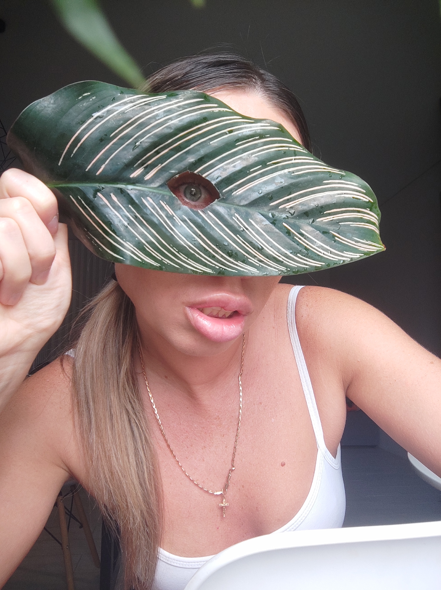 A woman with light skin, brown hair pulled back in a ponytail, and wearing a white tank top and gold necklace is holding a large, green leaf with white stripes in front of her face. A hole has been made in the leaf to reveal her left eye. The woman's lips are slightly parted. The background is blurred and partially obscured by the leaf. It is unclear whether this photo was taken in Votkinsk, Russia, or anywhere else.