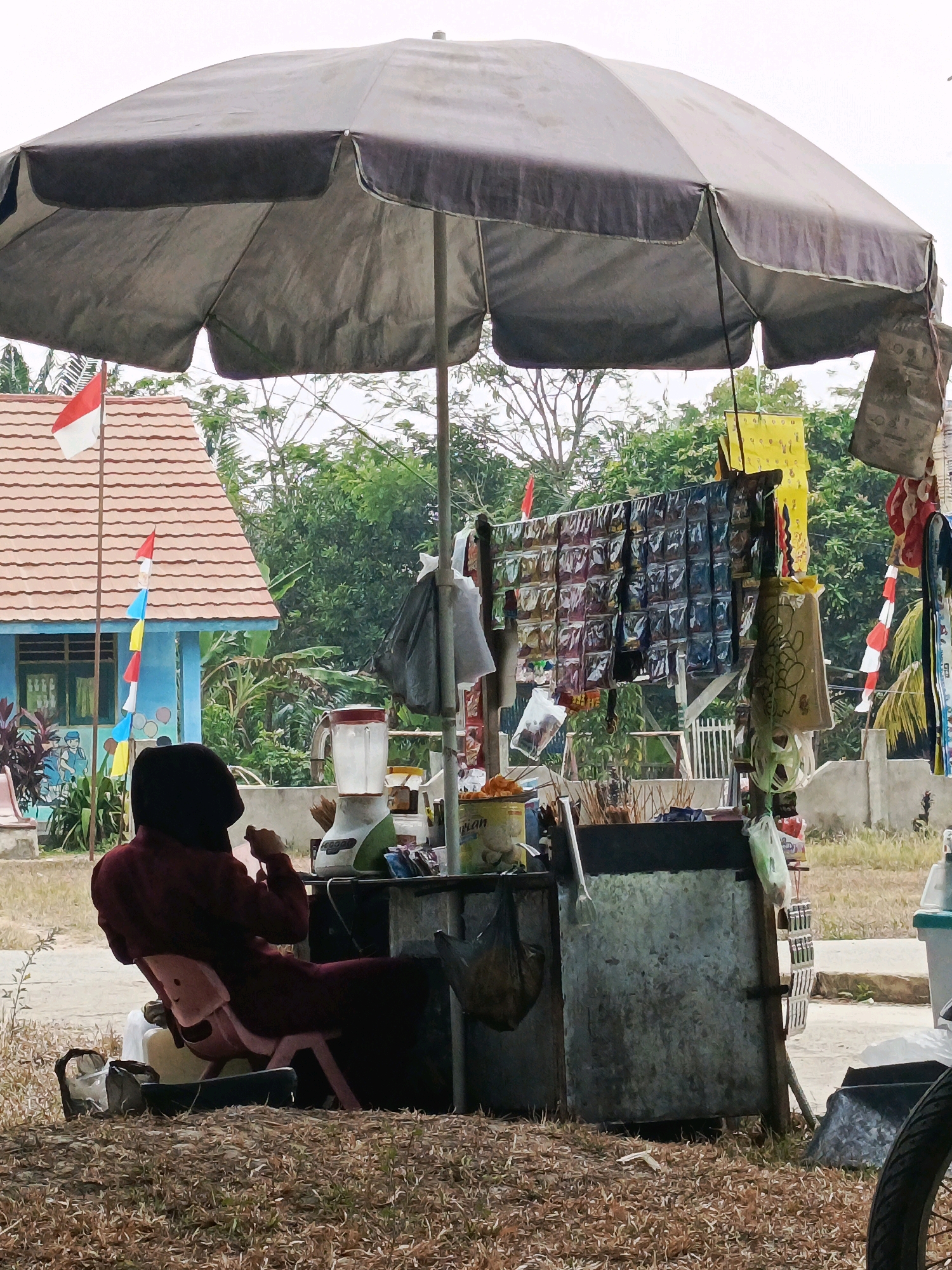 The image depicts a roadside stall under a large gray umbrella in Megang Sakti V, Indonesia. A person wearing a maroon headscarf and cloak is seated in a pink plastic chair facing a stall. The stall is equipped with a blender, a metal container, and various items including what appear to be packaged snacks or goods displayed on a rack. Behind the stall, there is a blue building with a red-tiled roof, adorned with Indonesian flags. The environment consists of dry grass and vegetation, with trees in the background. The overall scene suggests a humble commercial setup operating outdoors.