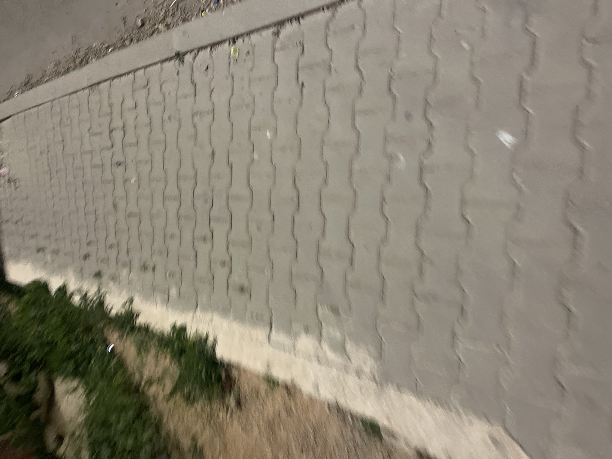 A ground-level, oblique angle image depicts a paved area consisting of interlocking concrete blocks. A strip of green vegetation borders the paved area on the left, transitioning into a patch of dry, brown earth. A concrete curb runs along the top of the paved area. The image appears to be taken at night or in low light conditions. The exact location within the city of Ouaouizert, Morocco, cannot be determined without further information.