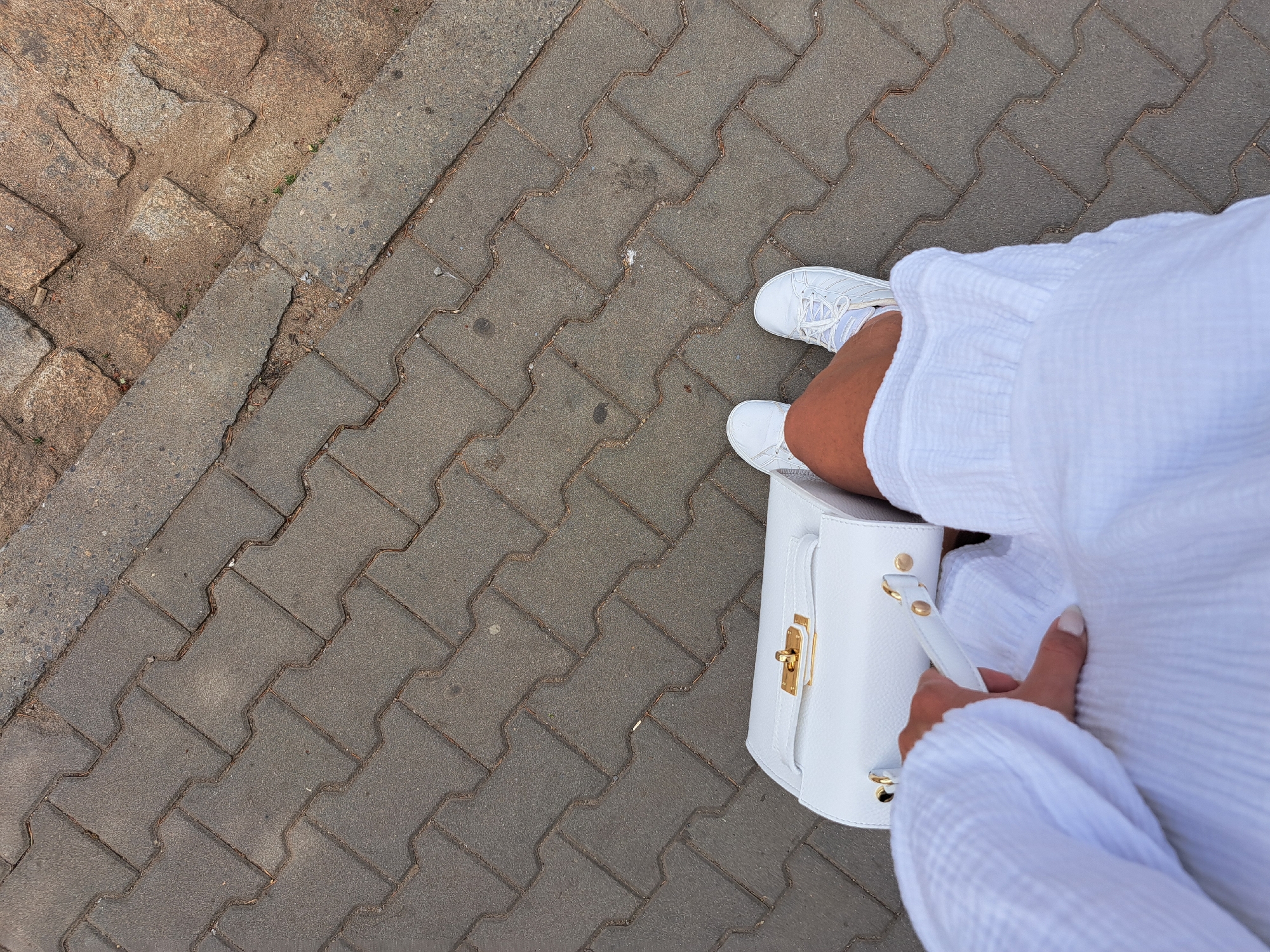 In a top-down view of a paved walkway, a person is captured mid-stride. They are wearing white sneakers and a white robe, and are holding a white handbag with a gold clasp. The walkway is constructed of interlocking grey pavers, with a strip of reddish-brown cobblestones bordering the right side of the frame. The scene appears to be an outdoor urban environment.